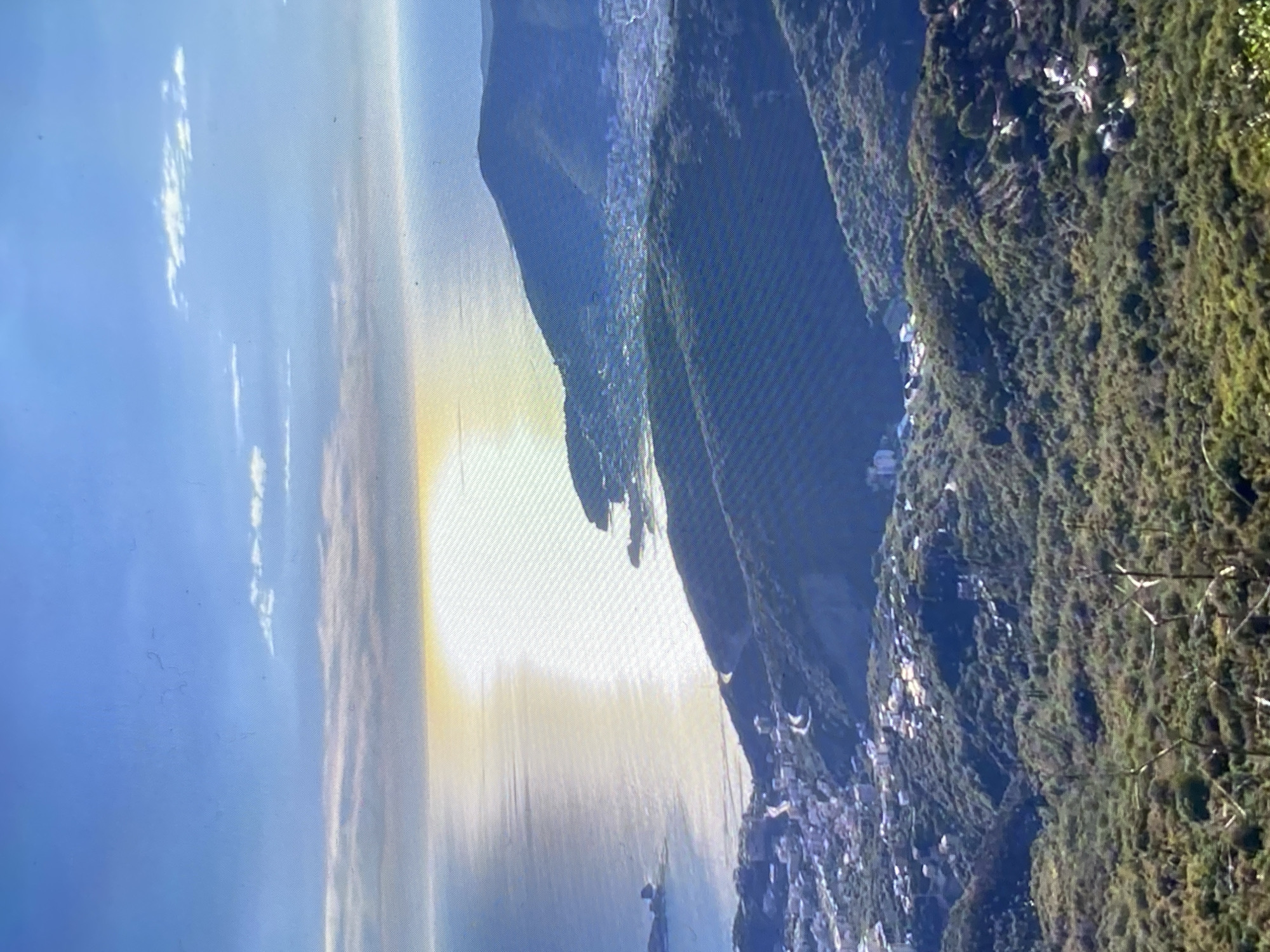 An aerial perspective captures a sun-drenched coastline with rolling green hills covered in trees and dotted with buildings. The sun is low on the horizon, casting a bright, shimmering reflection across the calm blue sea. A small town with white buildings is nestled in the valley between two hills, and a road winds its way through the landscape. The sky is clear blue with a few wispy clouds.