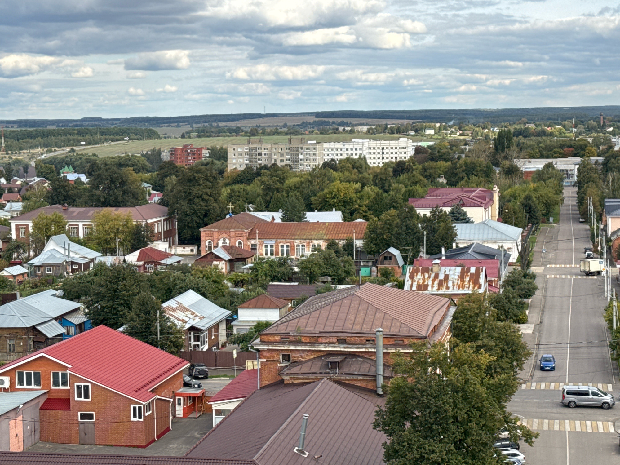 The image depicts a panoramic view of a city, identified as Zaraysk, Russia, from an elevated vantage point. The cityscape is characterized by a mix of residential buildings, some with red tile roofs and brick facades, alongside larger, multi-story concrete structures visible in the distance. A prominent feature is a tree-lined street that winds through the town, with several vehicles, including cars and a truck, navigating its lanes and pedestrian crossings. The surrounding environment is verdant, with extensive tree cover interspersing the urban architecture, and the distant landscape shows rolling hills and fields under a cloudy sky. The overall impression is of a well-established, mid-sized town with a blend of older and more modern architectural styles, set within a rural context.