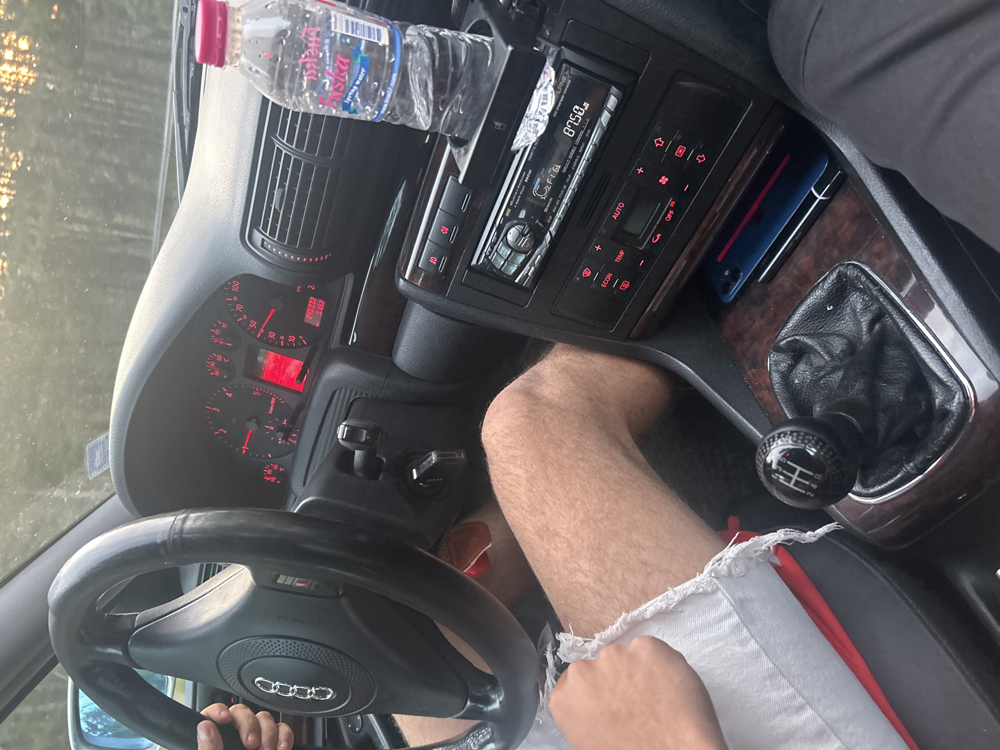 The image captures a partial view of a car's interior, likely an Audi due to the visible logo on the steering wheel and an "S line" badge. The perspective is from the passenger side, looking towards the driver and the dashboard. The driver's hand is on the steering wheel, and their bare legs, wearing white frayed shorts with a red lining, are visible.
The dashboard is illuminated with red lights, displaying analog gauges for speed, RPM, fuel level, and engine temperature. A digital readout shows an odometer reading of 248,832 km, a trip meter of 519.2, and an outside temperature of 19.5°C. An aftermarket Alpine brand car stereo is installed in the center console, displaying "87.50" MHz (likely an FM radio frequency) and "F16". Below it, the climate control panel features buttons such as "AUTO", "TEMP", "ECON", and "OFF".
A bottle of "SKA Spring water" with the website "www.lina07.com" printed on its label is placed in a cup holder. The car features dark wooden trim on the center console. A manual gear shifter with a carbon fiber-look knob clearly shows the 5-speed and reverse (1, 2, 3, 4, 5, R) pattern. Tucked next to the shifter are a smartphone in a blue case and what appears to be a vape pen. Golden sunlight streams through the windshield, indicating either early morning or late afternoon, suggesting the car is outdoors in a sunny environment, consistent with the provided location context of Dzhebel, Bulgaria. The high mileage suggests a well-used vehicle, likely an older model Audi.