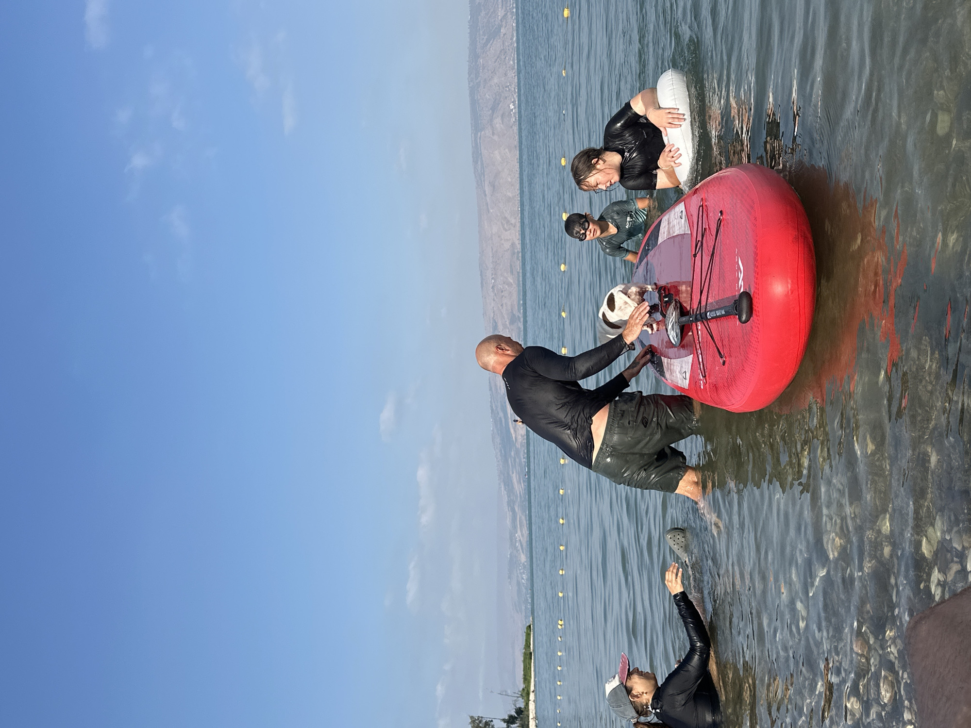 In the city of Tiberias, Israel, on a clear, sunny day, a group of people and a dog are in the shallow waters of the Sea of Galilee. A man in a black rash guard and dark swim shorts is kneeling on a red paddleboard, reaching down to a white and brown dog standing on the board. To the left of the paddleboard, a woman wearing a black rash guard and a baseball cap is partially submerged in the water, reaching out towards the paddleboard. To the right of the paddleboard, two children are also in the water. One child, wearing a dark green rash guard and swim goggles, is looking towards the paddleboard. The other child, wearing a black rash guard, is holding onto a white inflatable ring. The paddleboard is positioned in the center of the frame, with its front end closer to the viewer. The water is clear, with visible pebbles and stones on the seabed. In the background, a distant shoreline with hills and vegetation can be seen under a vast blue sky with a few scattered clouds. Yellow buoys are floating on the water's surface, marking a boundary.