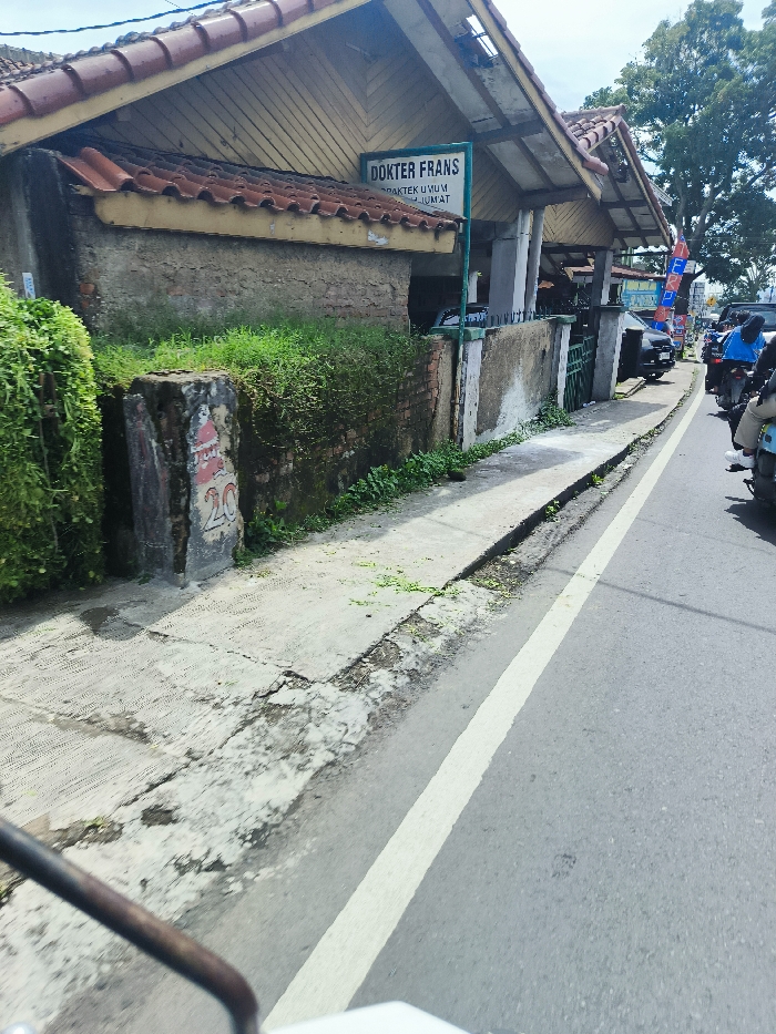 The image shows a narrow street in Ciwidey, Indonesia, with a concrete sidewalk on the left and a paved road on the right. A white line separates the sidewalk from the road. The sidewalk is overgrown with green vegetation and there is a brick wall on the left side of the image. A sign that says "Dokter Frans Praktek Umum Jumat" is visible on the right side of the image. There is a building on the right side of the image, which is likely a medical clinic. The image is taken from a low angle, looking down at the street from the perspective of someone on a motorcycle. There are several motorcycles parked on the right side of the road.