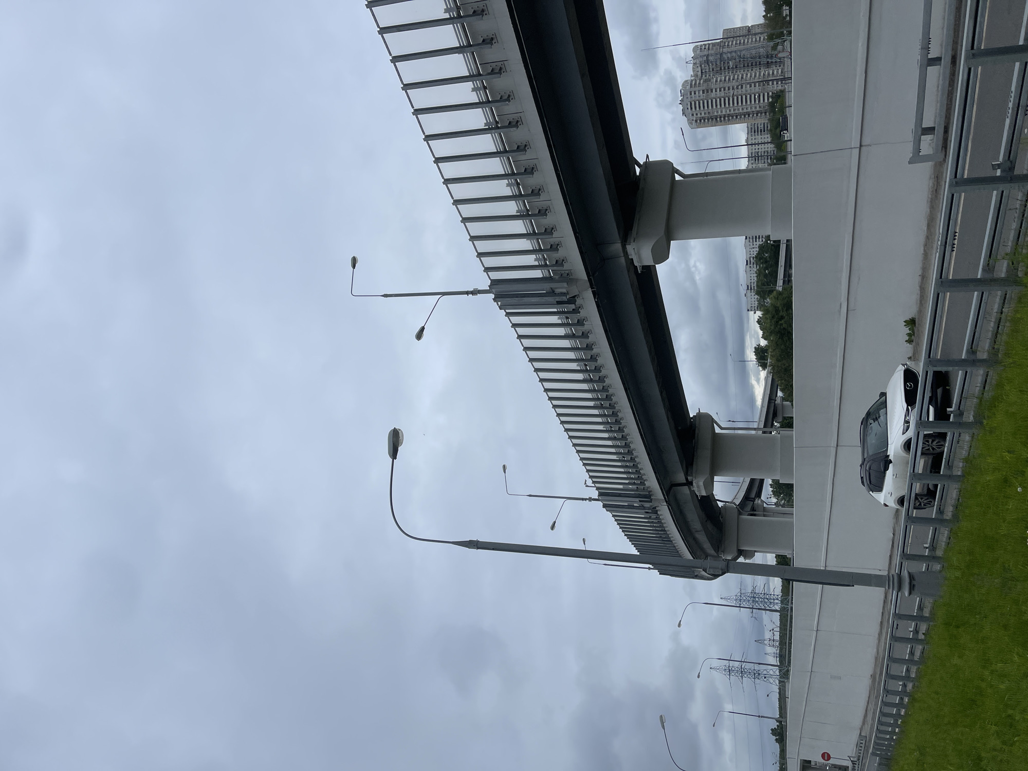 This image depicts a segment of the Moscow High-Speed Diameter (MSD) elevated highway in Moscow, Russia. The focus is on the structure of the highway itself, featuring a repeating pattern of steel supports and a concrete roadway. Street lamps are visible along the side of the road. In the background, there are high-rise buildings and power lines. A white car is parked on the side of the road next to a grass verge. The sky is overcast.