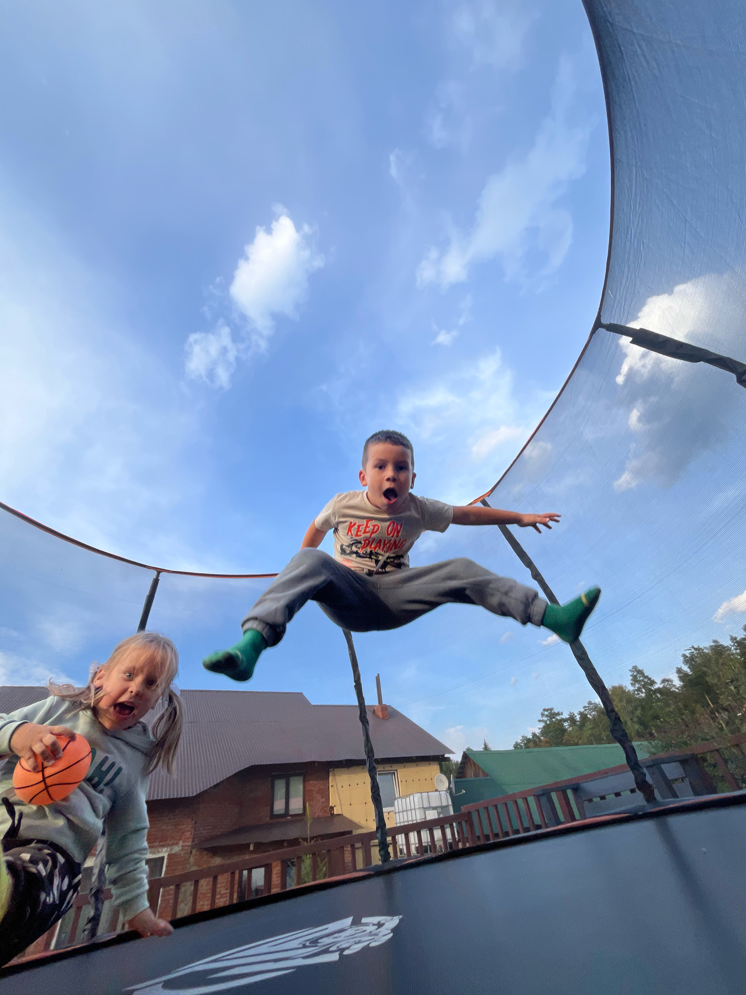 The image shows two children, a boy and a girl, jumping on a trampoline outdoors. The boy, positioned in the center of the frame, is mid-air with his legs spread and his mouth open in an expression of surprise or excitement. He is wearing a light-colored t-shirt with red lettering and grey sweatpants, along with green socks. To the left of the frame, the girl is also jumping, holding a small orange basketball in her hand. She has blonde hair in pigtails and is wearing a light-colored hoodie and patterned leggings. Her mouth is also open, suggesting she is making a sound or shouting.
The trampoline's safety net is visible, creating a curved frame around the upper part of the image, against a bright blue sky with scattered white clouds. In the background, a residential area with houses and trees is visible. The houses have dark, sloping roofs, and one appears to be under construction or renovation. A wooden fence is also present in the background. The overall setting suggests a suburban or rural environment. Based on the provided context, this scene is located in the city of Chaikovsky, Russia.