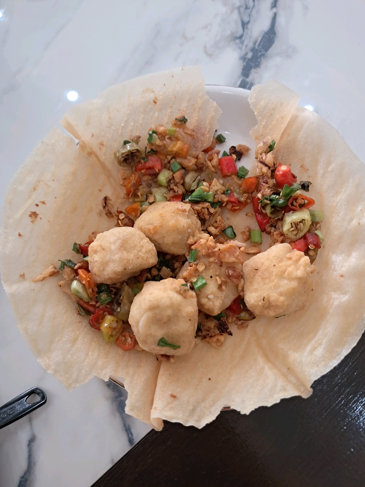 A close-up overhead shot shows a white plate filled with a serving of what appears to be deep-fried tofu with a stir-fried vegetable mixture and a crispy wrap. The tofu is light brown and irregularly shaped, with a slightly bumpy texture. The vegetable mixture is a colorful medley of chopped red chili peppers, green chili peppers, green onions, and what looks like minced meat or finely chopped nuts. The crispy wrap, made of a thin, translucent dough, is folded and serves as a base for the food, with its edges extending upwards and outwards. The food is placed on a white, marble-patterned surface, and a dark, textured surface, possibly a placemat or table, is visible in the lower right corner. A small, black metal utensil handle is visible on the left side of the frame. The lighting suggests an indoor setting with overhead illumination. The image does not contain enough information to determine the specific location in the city of Mempawah, Indonesia.