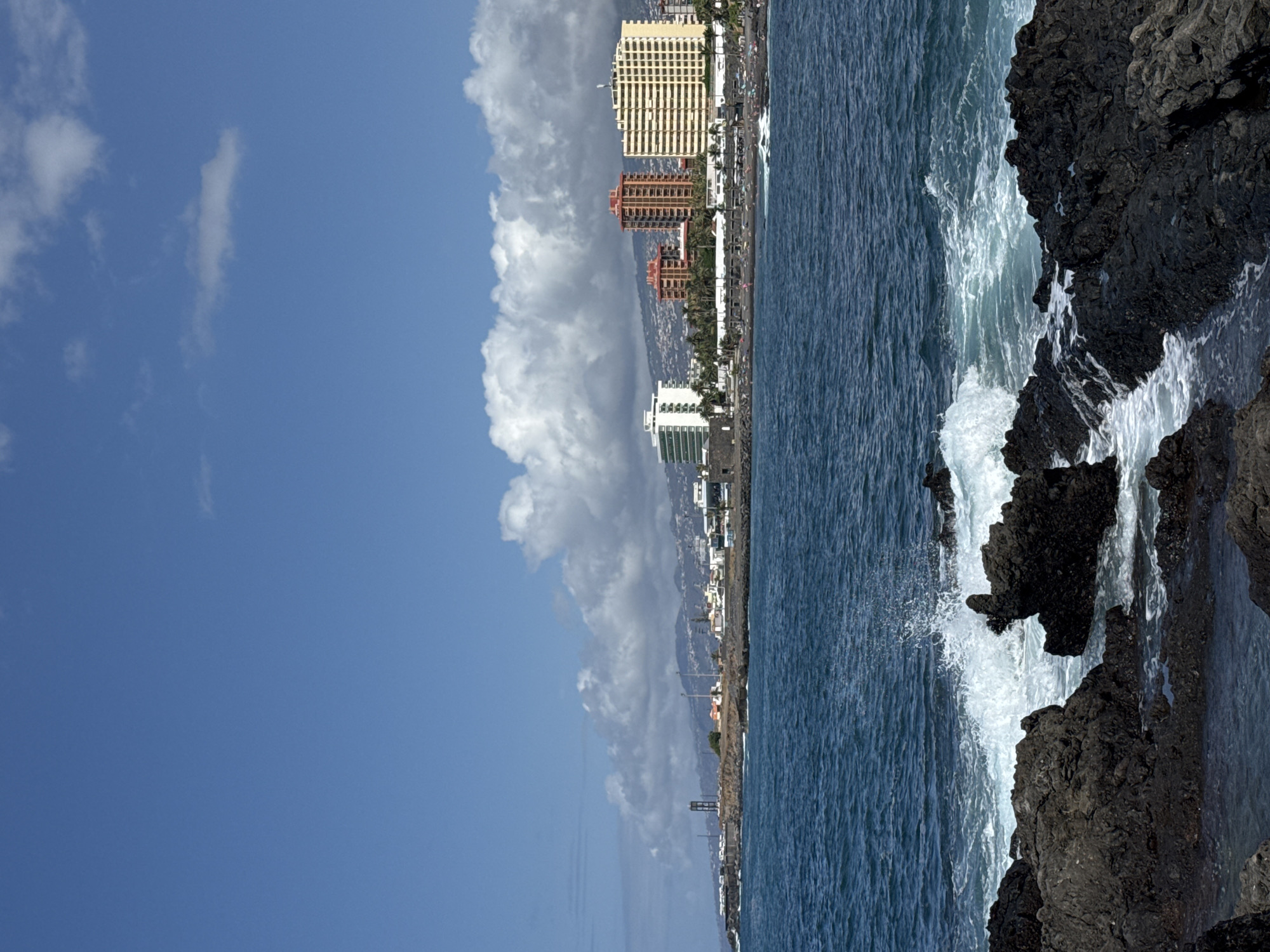 The image depicts a coastal scene with dark, volcanic rocks in the foreground, on which waves are crashing and creating whitewater. Beyond the rocks, a body of deep blue water stretches towards a coastline lined with buildings, palm trees, and a beach populated with many people. A dark tower stands on the left side of the coastline. Further inland, mountains are visible under a sky with scattered white clouds. The buildings on the coastline include a tall white structure on the left, followed by several red-toned buildings, and a large yellow building on the right. The overall scene suggests a sunny day at a coastal resort or city.