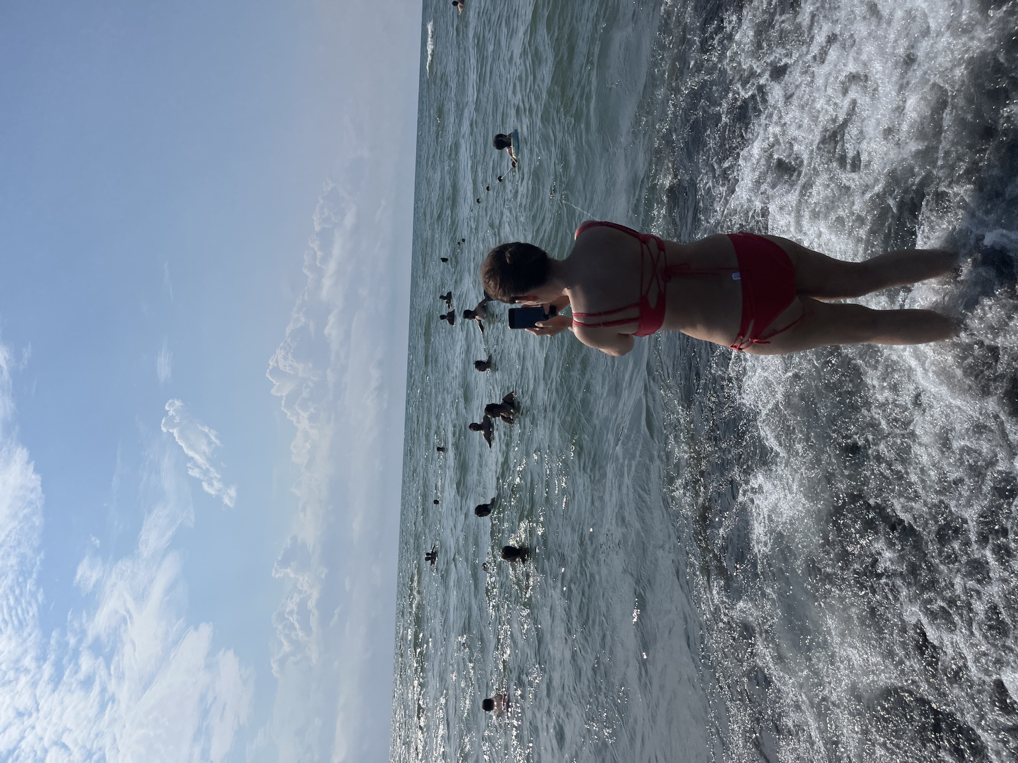 A medium shot captures a woman in a red bikini standing at the edge of the Black Sea in Ureki, Georgia, facing away from the camera and holding a smartphone to take a picture. The sea is filled with people swimming and floating, with some visible buoys or ropes in the water. The sky is bright blue with scattered white clouds. The water is choppy with white foam near the shore.