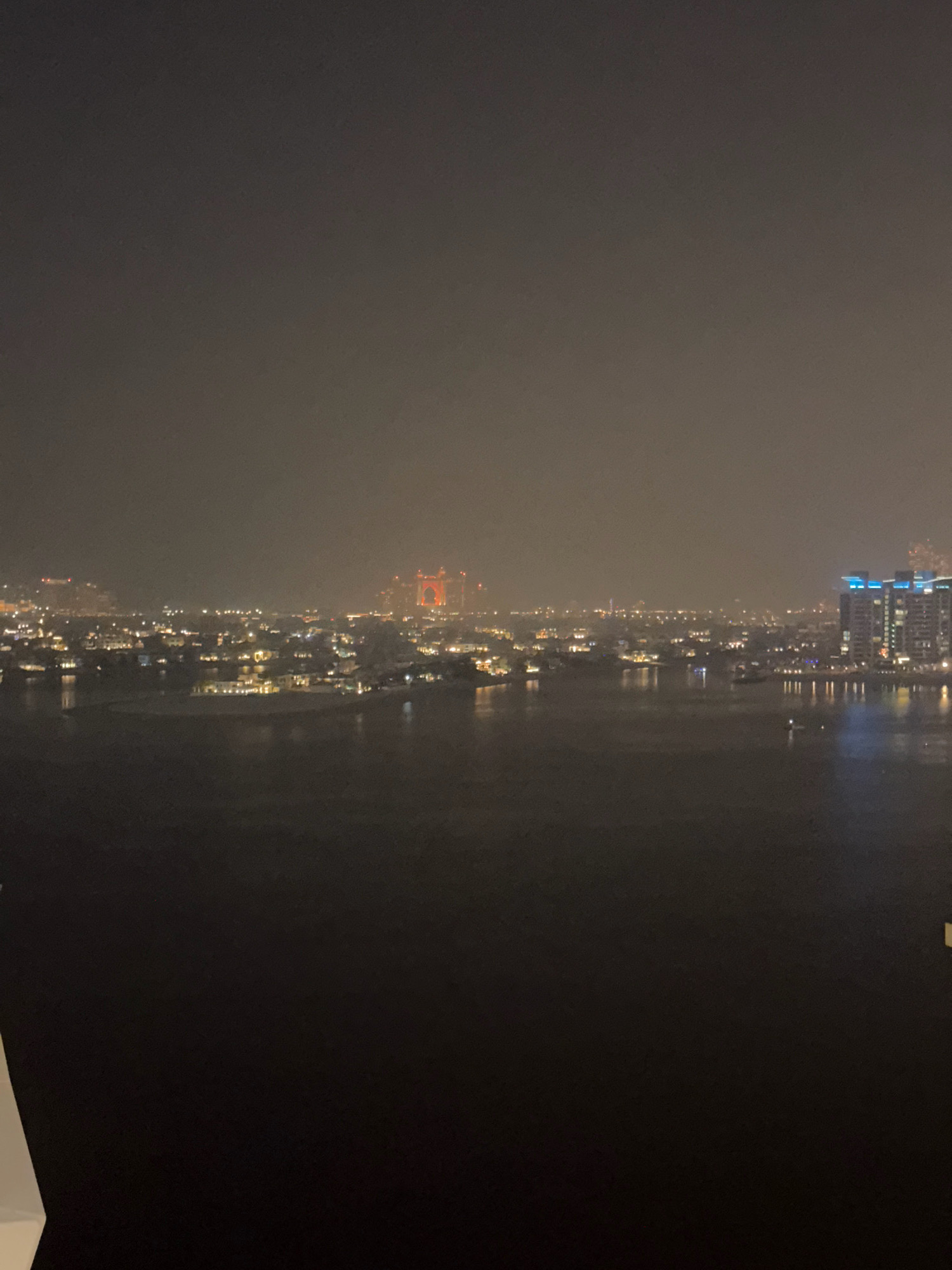 The image captures a nocturnal, hazy cityscape with illuminated buildings reflecting on the water. The scene is located in Dubai, United Arab Emirates, and appears to be taken from a high vantage point, possibly a hotel room or apartment.
In the foreground, a dark body of water dominates the lower half of the image. The surface of the water is mostly calm, with faint reflections of distant lights.
The middle ground reveals a sprawling urban area with numerous buildings of varying heights. Many structures are lit from within, creating a warm glow that pierces the atmospheric haze. The lights are concentrated, suggesting a densely populated and developed district. A prominent feature in the mid-distance is a large building with distinctive orange-red lighting, possibly a hotel or entertainment complex. To the right, a modern skyscraper with blue-lit vertical accents stands out against the darker sky.
The background is characterized by a thick, pervasive haze that softens the outlines of distant structures and diffuses the light. The sky above is a muted, dark gray, with no visible stars or moon.
There are no people or clear signs of activity depicted in the image. The overall impression is one of a quiet, illuminated urban environment at night, viewed through a veil of atmospheric conditions. The architecture suggests a modern, affluent city.