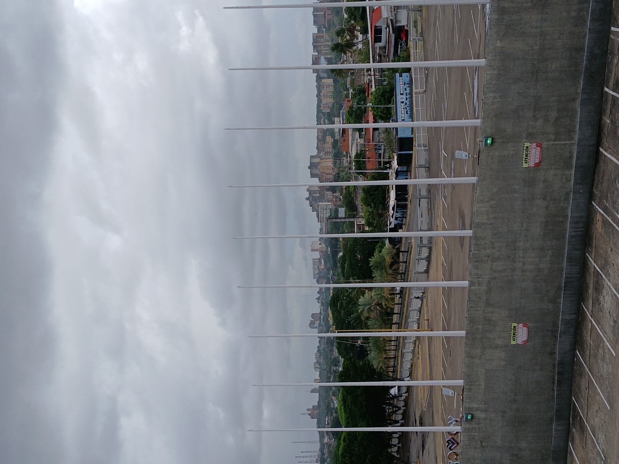 The image depicts an elevated view of an urban landscape under a cloudy sky. The foreground features a concrete wall with parking space markers. Along the wall are several tall, white poles extending vertically. Behind the poles, there is an open parking lot with painted lines marking parking spaces. Beyond the parking lot is a view of a city with buildings of varying heights and architectural styles. Patches of green foliage are interspersed throughout the cityscape. Signs in Spanish are seen on the wall.