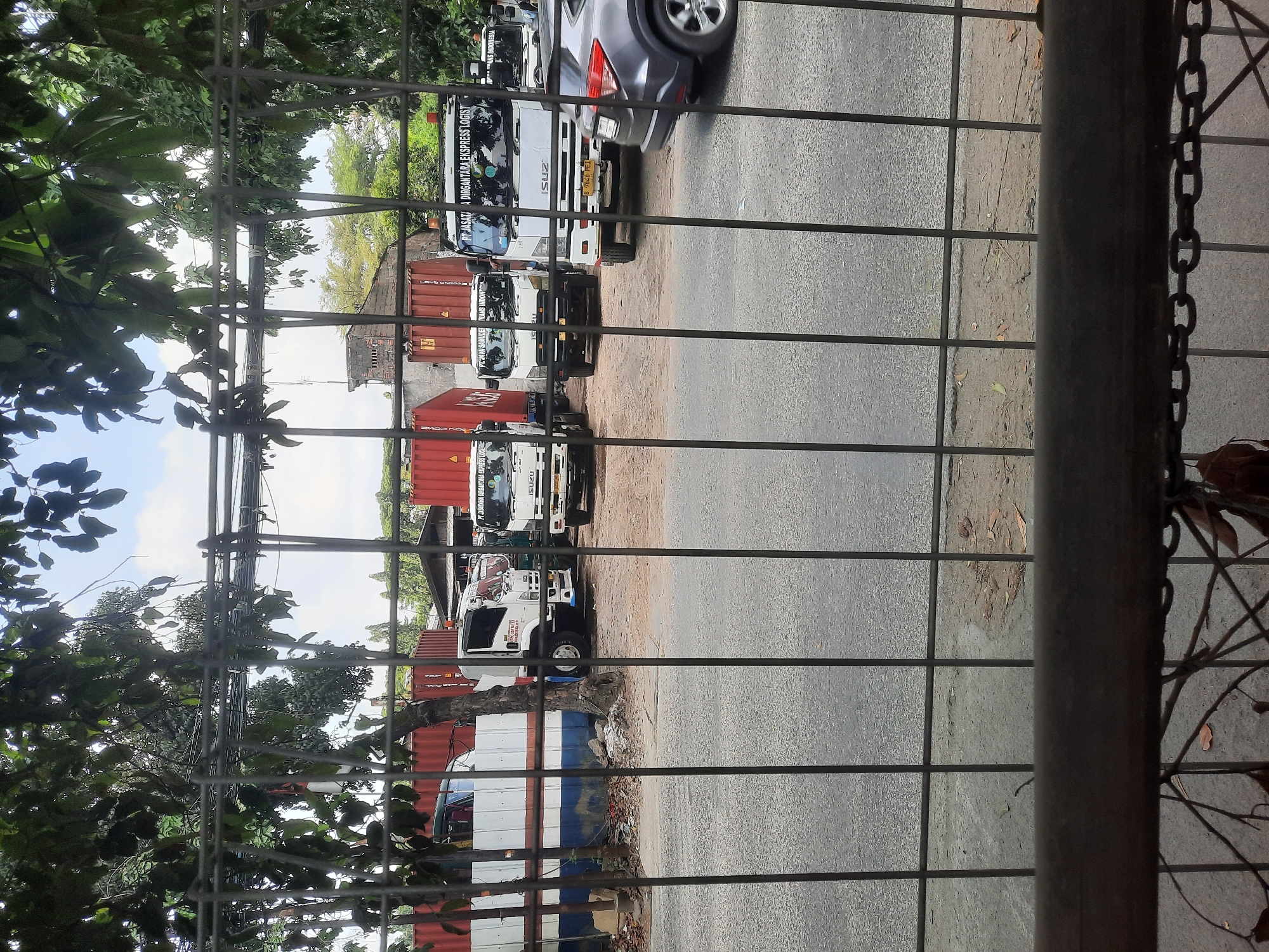The image shows a row of trucks parked on a dirt lot behind a metal fence. The trucks are white and have the logo "PT. JASA DIRGANTARA EKSPRESS LOGIST" on them. Some of the trucks have shipping containers loaded on them. In the background, there are some buildings and trees. A grey car is also visible on the right side of the image. The scene appears to be in an industrial area in North Jakarta, Indonesia.