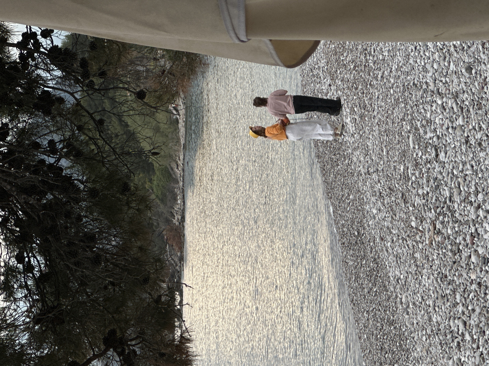 The image captures a beach scene in Bar, Montenegro. The foreground features a rocky beach composed of numerous small, rounded stones. Adjacent to the beach is the shimmering water surface, reflecting light and creating a rippled texture. On the beach, two people stand. One individual wears a light pink top and dark pants, while the other sports an orange top and white pants, accented with a yellow hat. They appear to be interacting, possibly conversing, with their backs turned towards the vantage point. The left side of the frame is dominated by the dark silhouette of a tree, laden with pine cones. The branches extend into the scene, framing the view. The top of the image reveals a partial view of a light-colored awning or shelter.