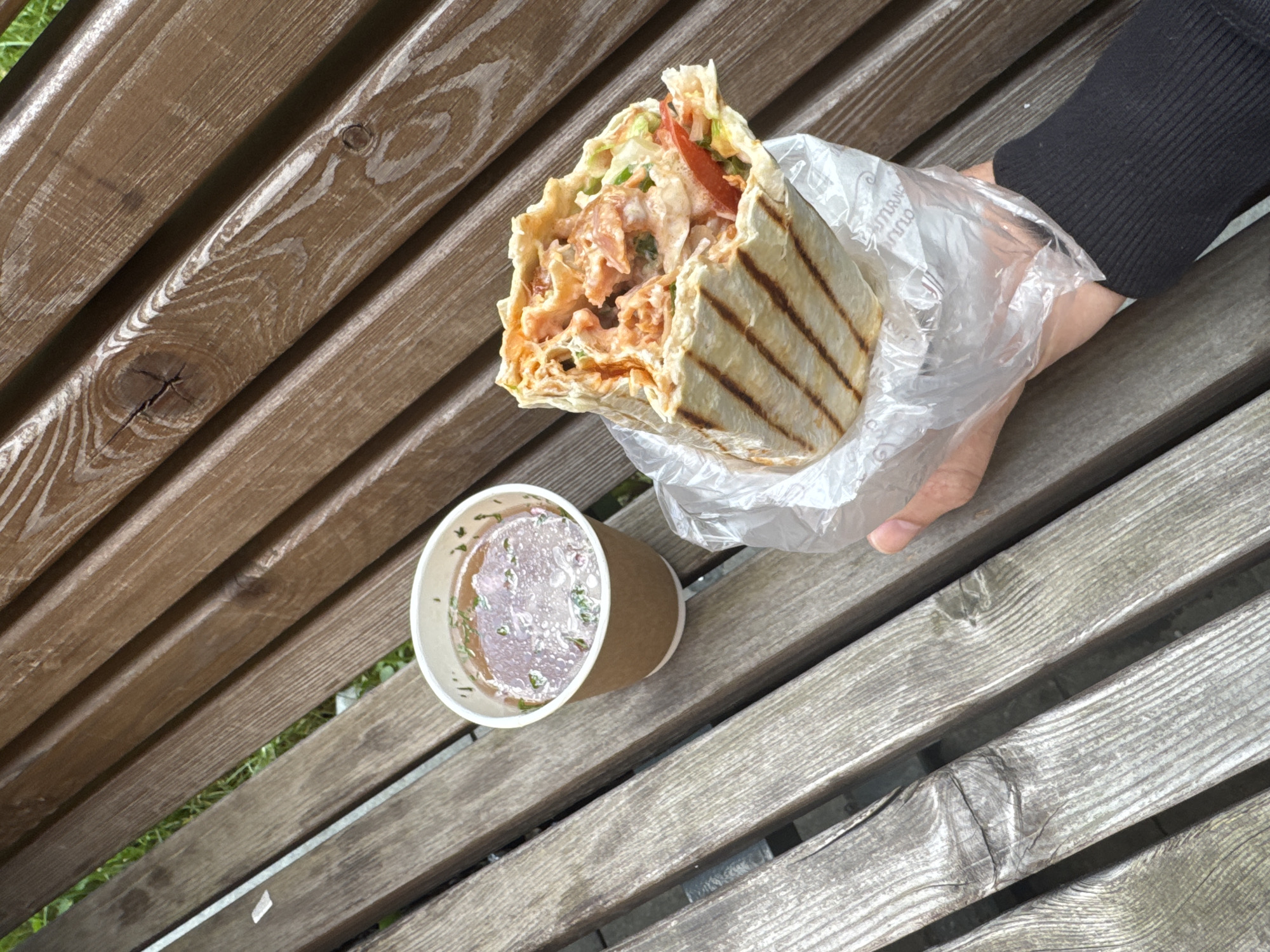 The image shows a person holding a shawarma in a plastic bag over a wooden bench. Beside the shawarma is a small cup of liquid with green herbs. The bench is made of weathered wood, with visible grain and knots. The location is likely in Moscow, Russia, based on the style of the shawarma, which is a popular street food in the region. The person is wearing a dark-colored long-sleeved shirt. The scene appears to be outdoors.