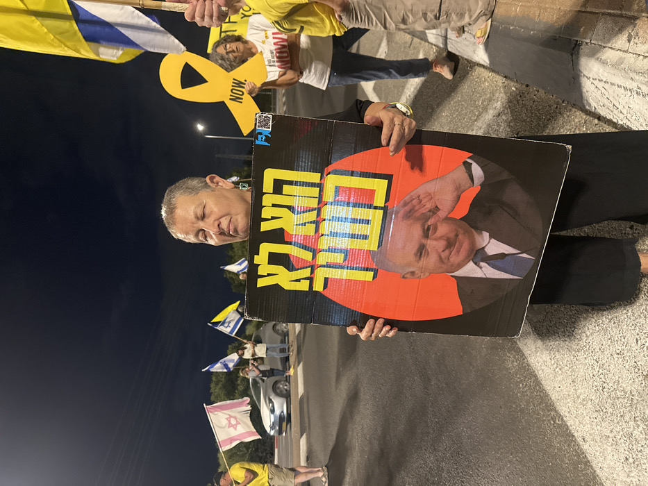 A middle-aged woman stands centrally at a nighttime protest in Mishmar Hasharon, Israel, holding a large, rectangular cardboard sign. She has short grey hair, wears dark clothing, and her eyes appear closed or heavily squinted.
The sign features a black background with yellow Hebrew text: "הוא לא" (Hu lo - He is not) at the top, and "במשמר" (B'Mishmar - In Mishmar / A guard) below. Centered within a red circular motif is an image of Benny Gantz, smiling and performing a salute while wearing a suit. A small, indistinct QR code or logo is visible in the sign's upper right.
In the background, to the left, several individuals are present; one person in a yellow shirt holds a white flag with a pink Star of David, and others hold blue and white Israeli flags. A silver car is parked near the curb. To the right, another woman, wearing a white t-shirt with partially visible text "BRING THEM HOME NOW!", holds a large yellow ribbon cutout labeled "NOW.". An arm holding an Israeli flag is also visible on the right. Artificial lighting illuminates the asphalt road, where white markings are visible, and distant trees are silhouetted against the dark sky.