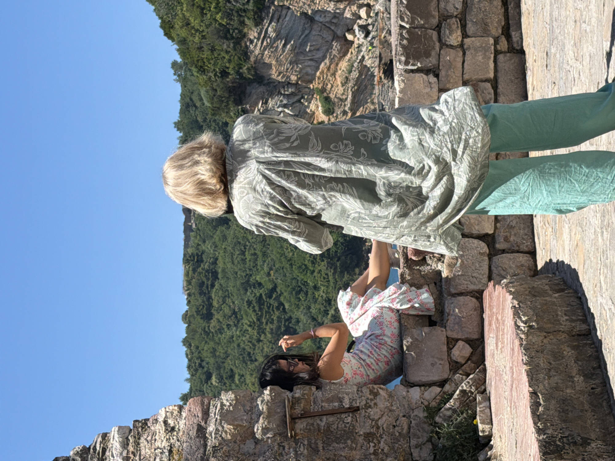 The image shows two women on a stone wall overlooking a rocky coastline with lush green vegetation. The woman in the foreground, viewed from behind, has blonde hair and is wearing a light green patterned shirt and light teal pants. She appears to be looking out at the scenery. The woman in the background is seated on the stone wall, facing away from the viewer and to the left. She has dark hair and is wearing a floral-patterned dress and sunglasses. She is touching her hair. The stone wall appears to be part of an old fortification, possibly in the vicinity of the Citadel of Budva or the walls of the Old Town of Budva, Montenegro, given the coastal rocky landscape and historical architecture. The sky is clear and blue, indicating a sunny day.