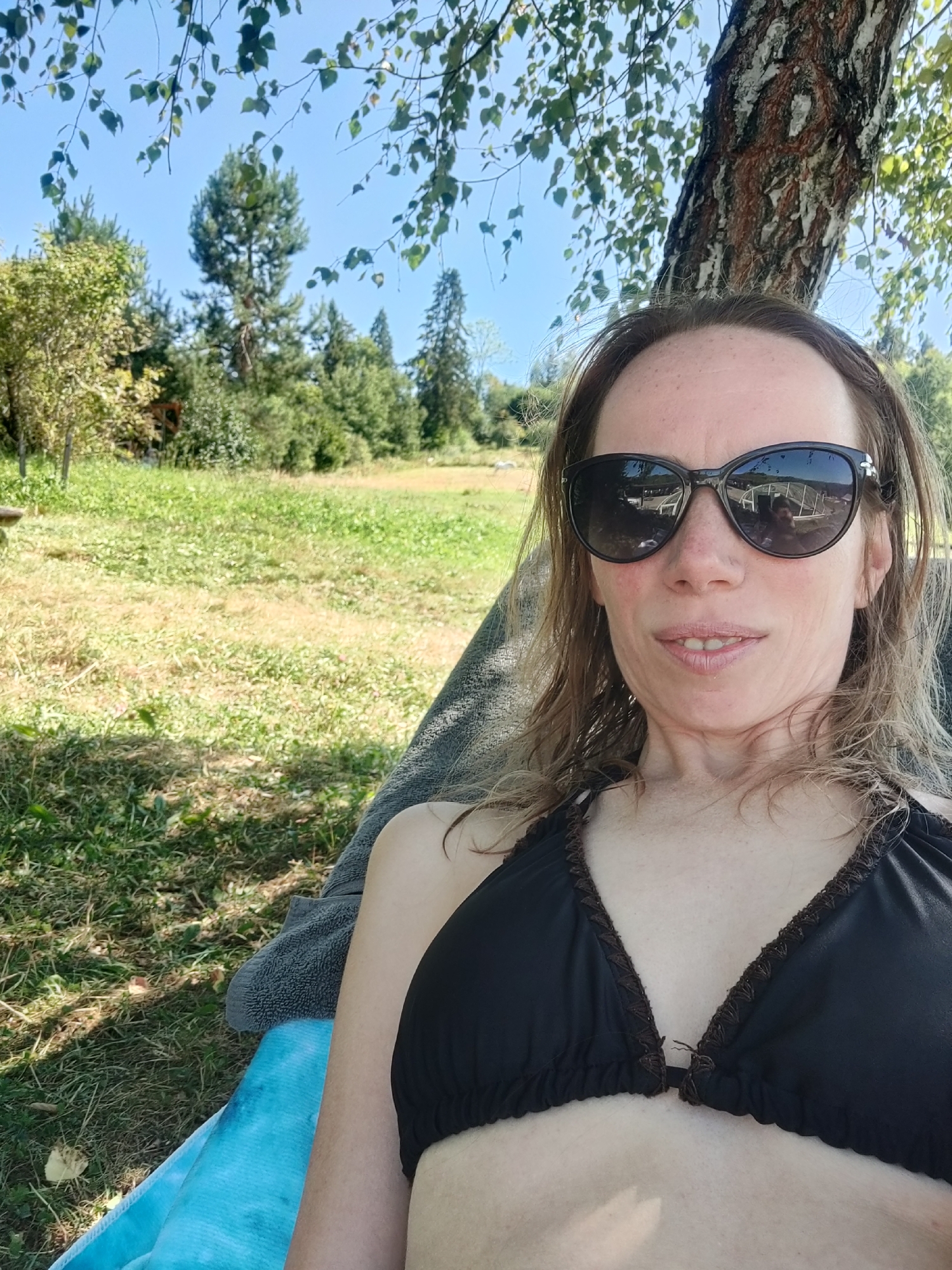 A woman with brown hair and sunglasses is lying down outdoors, possibly on a hammock or lounge chair, under a tree with dappled sunlight. She is wearing a black bikini top. The background shows a grassy field, trees, and a bright blue sky. The image appears to be a selfie taken during daytime. The location is not discernible from the image itself and cannot be determined to be Izky, Ukraine.