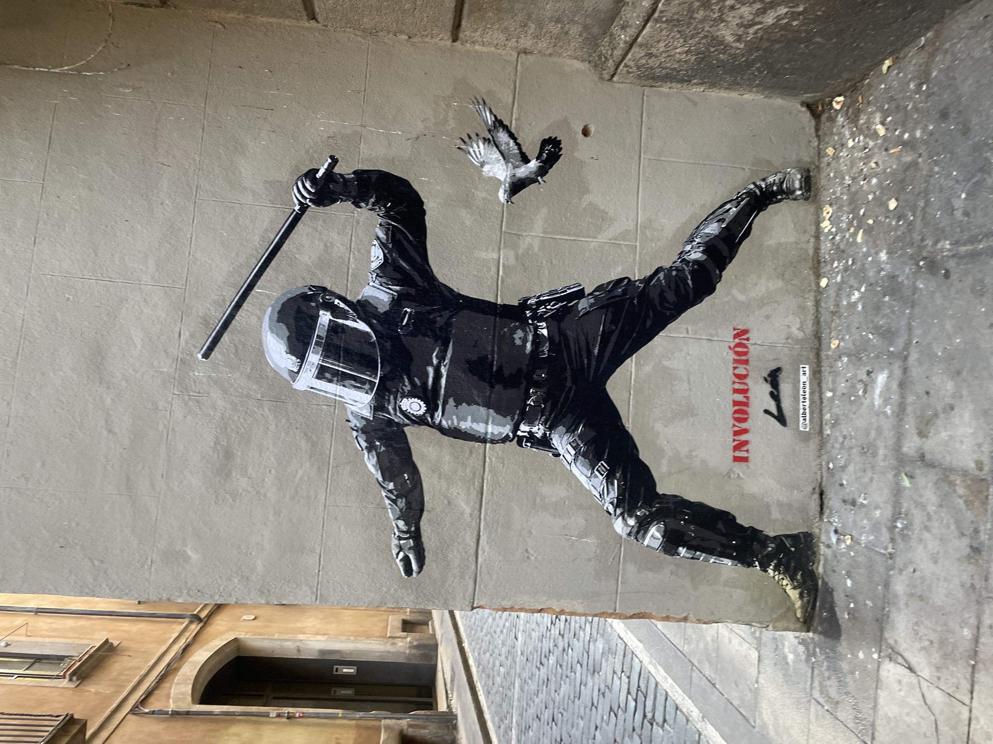 This image shows a street art mural on a textured, light-colored wall, located in Barcelona, Spain. The mural depicts a riot police officer in full gear, including a helmet with a visor, tactical clothing, knee pads, and boots, holding a baton raised as if to strike. Facing the officer, a dove is depicted in flight, with its wings spread. Below the mural, in red spray paint, is the word "INVOLUCIÓN", followed by a stylized signature and the handle "@alberteleon_art". The ground in front of the wall is paved with cobblestones and shows signs of wear and debris, suggesting an urban street setting. To the left of the mural, a section of an ochre-colored building with an arched doorway and a window is visible, with a narrow, paved street leading away from the viewer. The overall scene captures a moment of tension and symbolism between authority and freedom, rendered as street art in a public urban space.