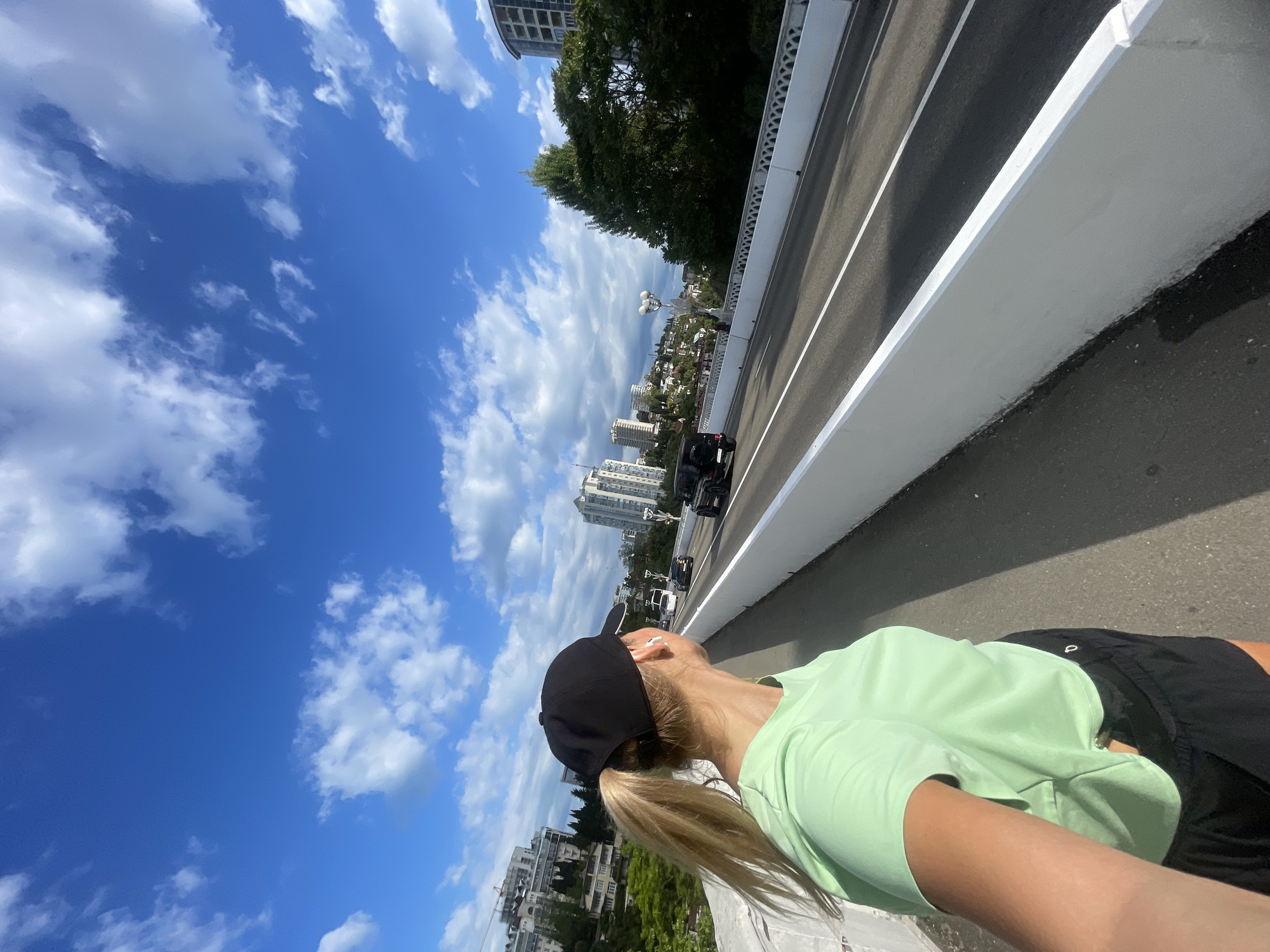The image shows a woman standing on a bridge, likely taking a selfie. She is wearing a light green shirt, black shorts, and a black baseball cap, with her blonde hair in a ponytail. Cars are visible on the bridge beside her. The background includes a blue sky with fluffy white clouds, trees, and buildings, indicating a city setting. Based on the architectural style and urban landscape, the location is possibly Sochi, Russia.