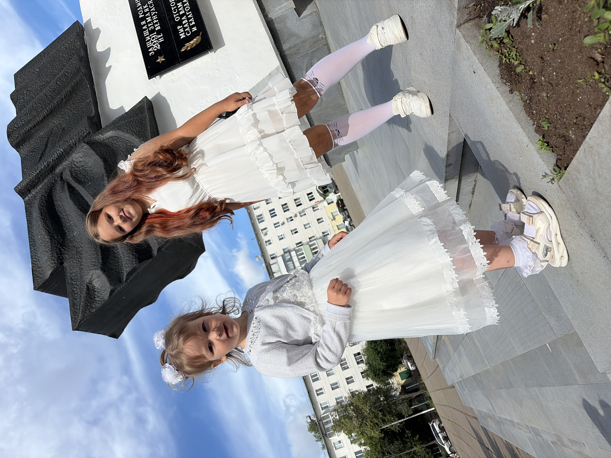 Two young girls, one younger and one older, stand on a concrete staircase in front of a memorial monument. The younger girl, on the left, wears a white, sparkly dress and a silver cardigan, with white bows in her hair. The older girl, on the right, wears a white tiered dress and white knee-high socks with cat faces on them. She has long, reddish-brown hair. Behind them, a large, dark, angular monument is visible, with a plaque attached to a white base. In the background, a modern, multi-story residential building and some trees are present. The sky is blue with scattered clouds. The memorial is located at the Victory Park in Chaikovsky, Russia, commemorating soldiers who died in World War II.