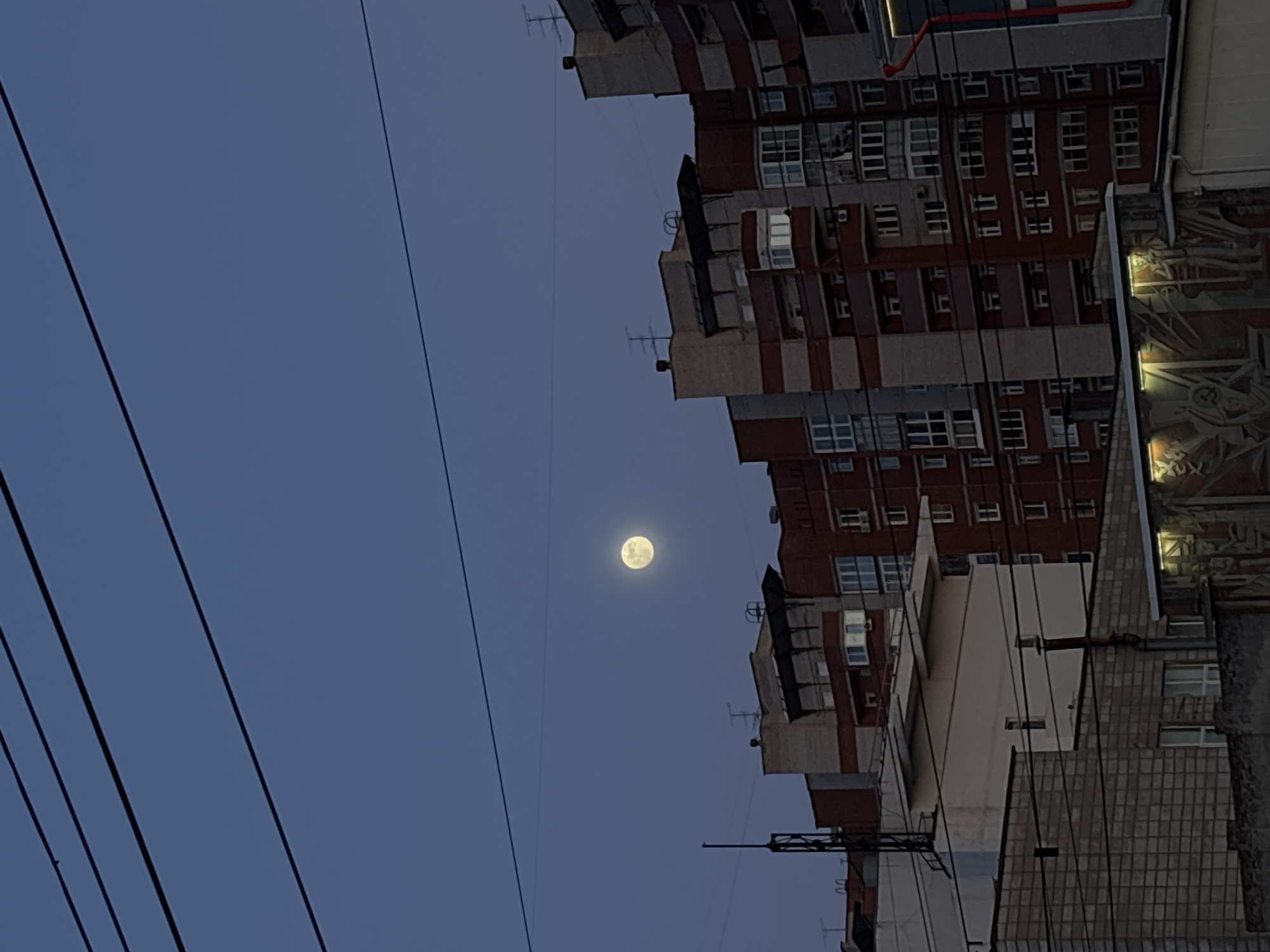 The image, taken in Perm, Russia, presents a nighttime urban scene. The focal point is the full moon, positioned in the upper middle of the frame against a deep blue sky crisscrossed by electrical wires. A multi-story brick apartment building dominates the right side of the image. The building is primarily clad in red brick with multiple rows of white-framed windows. Some windows are illuminated. A mural is visible on the adjacent structure. The image is rotated 90 degrees counterclockwise.