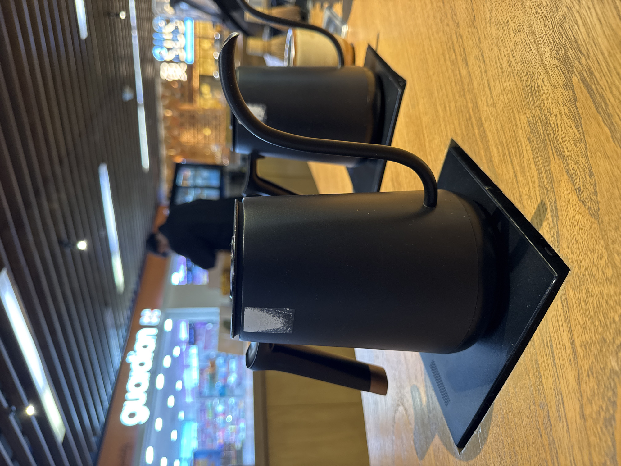 The image, originally rotated 90 degrees counter-clockwise, displays two black pour-over coffee kettles on a wooden surface, likely a table or countertop, in an indoor setting. The prominent kettle in the foreground is matte black with a distinctive gooseneck spout and a dark handle, which has a copper-colored accent at its end. This kettle rests on a square black base and has a partially peeled white sticker on its side. A second, similar black kettle is visible further back, also on a black base.
The background is out of focus, creating a bokeh effect from various light sources. To the left, there is a textured wall or paneling with vertical wooden slats and vertical light strips. To the right, a brightly lit store sign is visible, clearly showing the word "Guardian" in white text against an orange background, indicating a retail or shopping mall environment. Other blurred lights and shapes suggest the presence of more stores or displays. A person in dark attire can be vaguely seen in the deeper background. The scene depicts a product display, with no active event taking place. It is well-lit indoors, and the time of day or weather is not discernible.