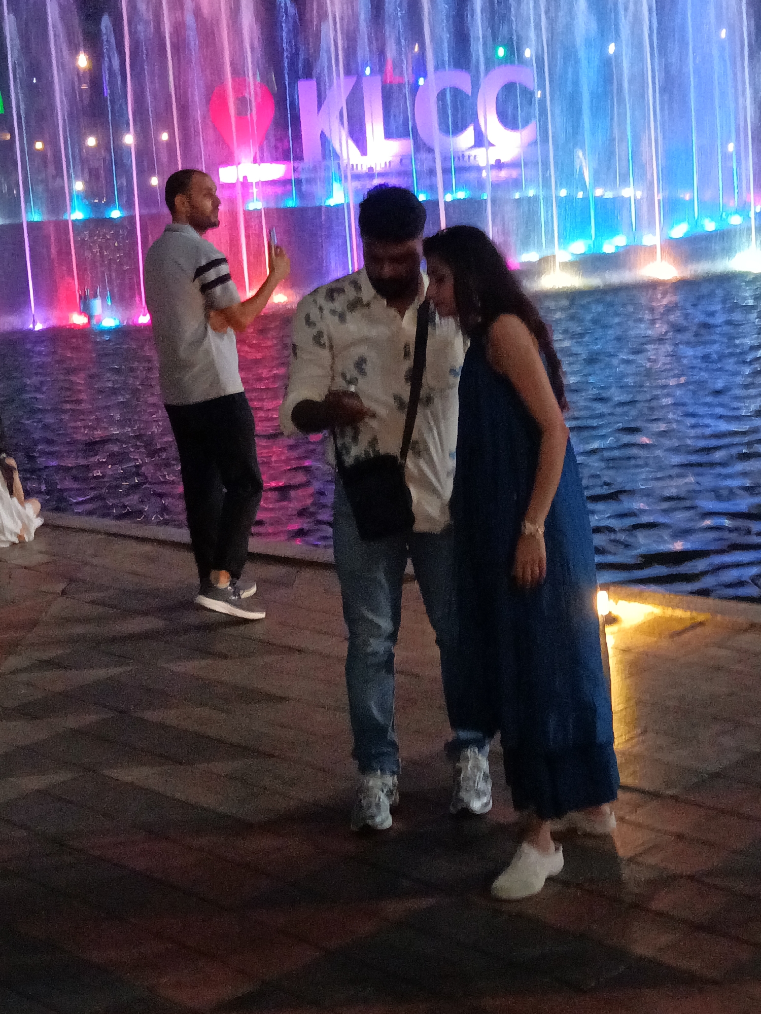 The image shows a group of people in front of the iconic KLCC fountain in Kuala Lumpur, Malaysia. A man in a light-colored shirt and dark pants is holding up a phone, seemingly taking a photo or video of the fountain. To his right, a couple stands close together, looking towards the fountain or at the phone. The man is wearing a patterned shirt and jeans, while the woman is in a long, dark blue dress. The fountain is illuminated with colorful lights, creating a vibrant and dynamic backdrop. The ground in the foreground is paved with dark tiles arranged in a patterned design, with shadows cast by the people and the fountain. The overall atmosphere is lively and festive, with the fountain's water jets creating a sense of movement and energy.