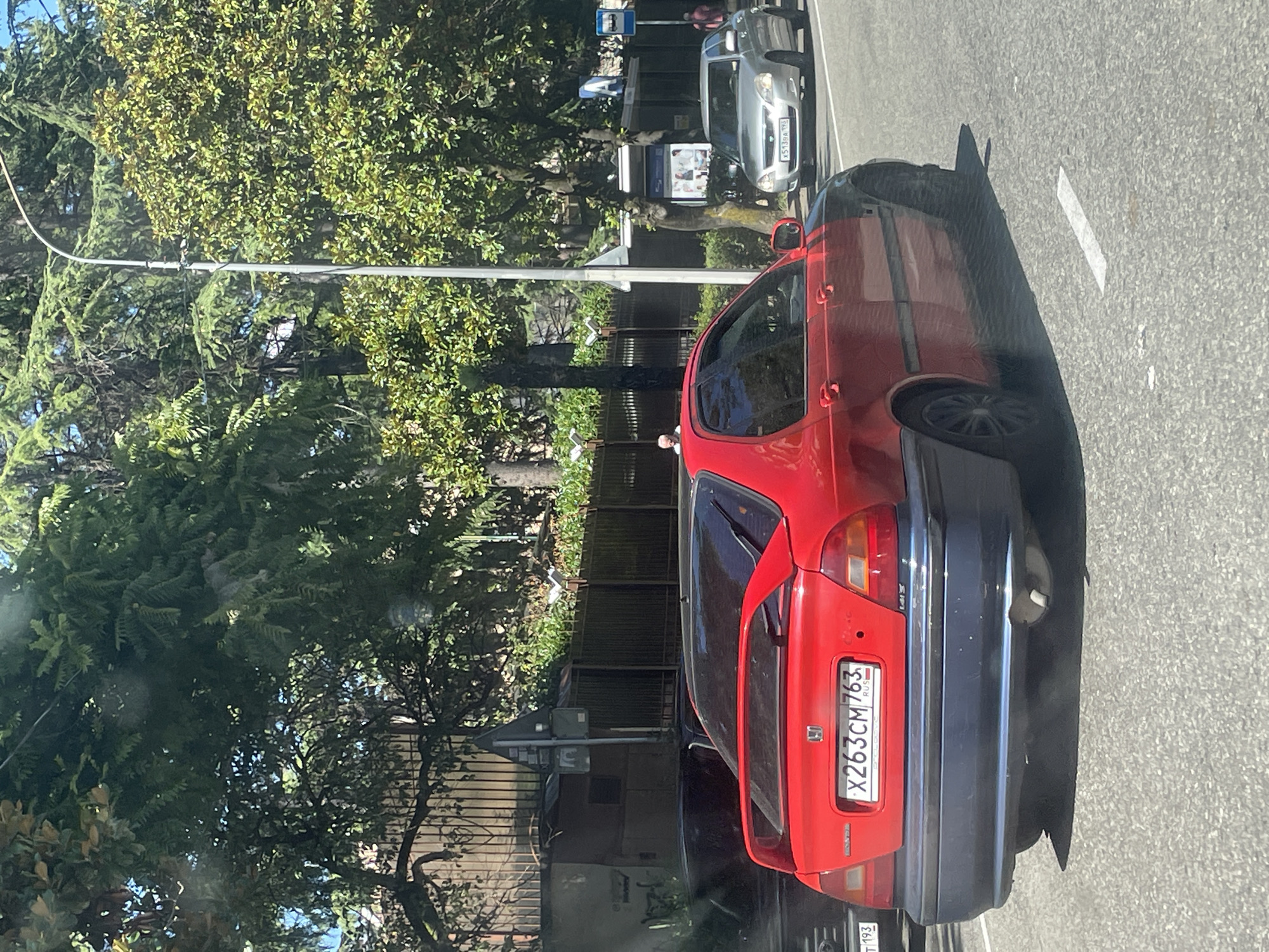A red Honda Civic is driving on a street in Sochi, Russia. The car has a spoiler on the back and a blue bumper. The license plate is X263CM763. A silver car is driving in the lane next to it, and a bus stop is visible in the background. There are trees and buildings along the side of the road. The weather is sunny.