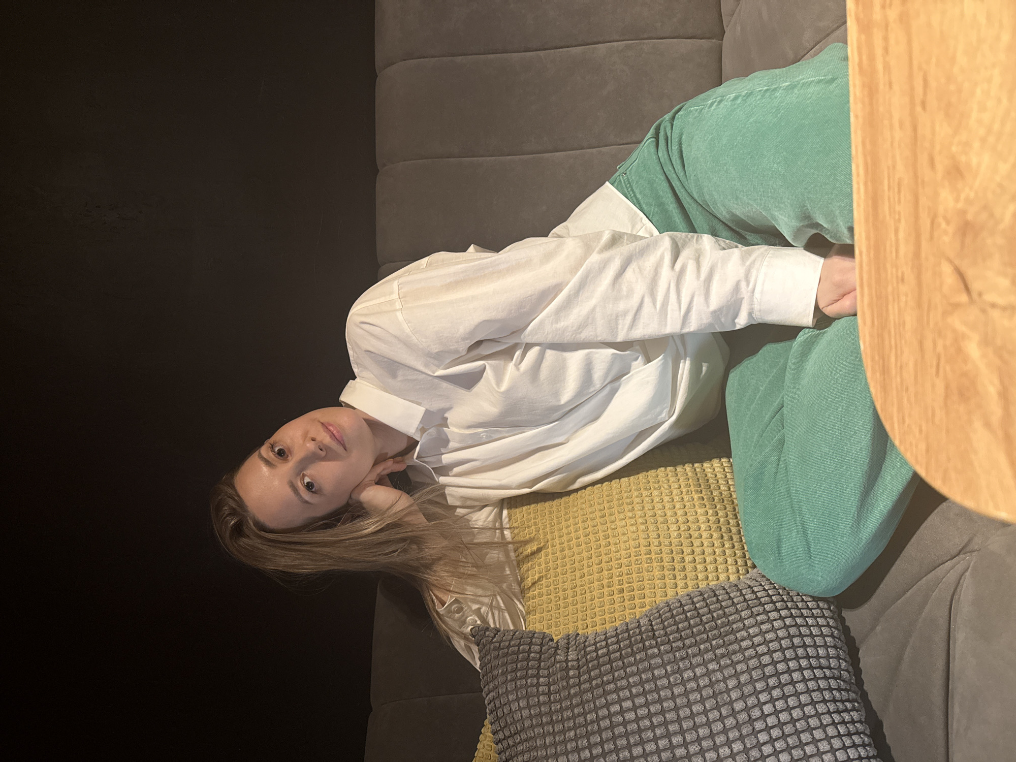 A woman is lying on a gray couch with a textured surface. She wears a white, button-down shirt and green pants. A textured gray pillow is placed under her head, and a yellow pillow is beside her. Part of a wooden surface is visible on the upper right side of the image. The background features a dark wall. The location is in Chaikovsky, Russia.