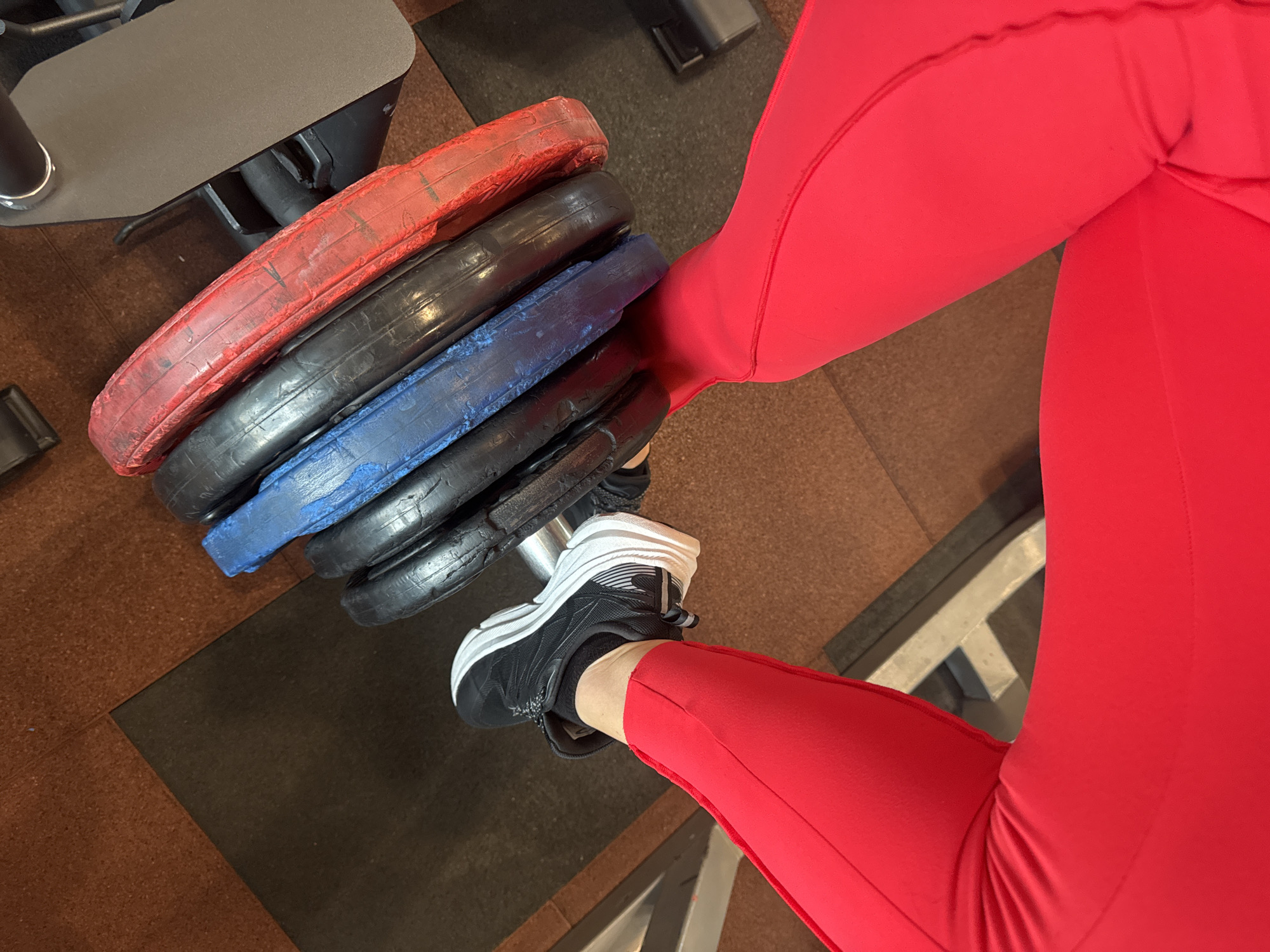 A high-angle, indoor shot shows a person wearing red leggings and black athletic shoes holding a stack of four weight plates. The top plate is red, followed by two black plates and a blue plate. The scene appears to be in a gym, evidenced by the weight plates and the presence of gym equipment partially visible in the background. The flooring consists of two different colors of rubber tiles. No information regarding the city of Vladikavkaz, Russia, is discernible from the image.