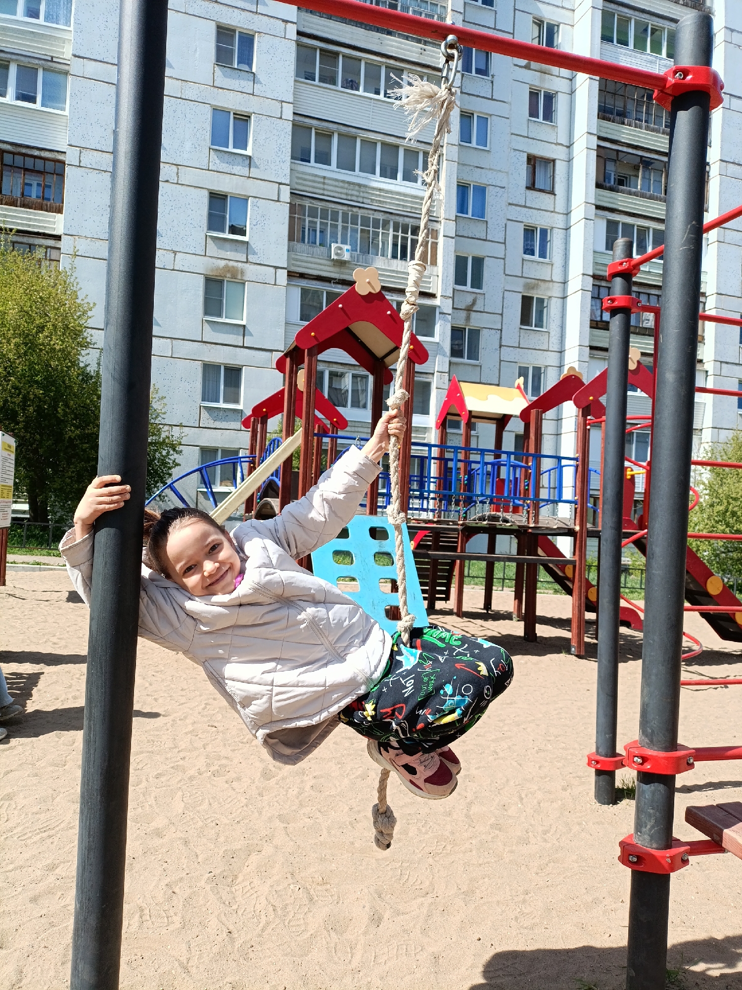 A young girl is hanging from a rope swing on a playground in Chaikovsky, Russia. The girl is wearing a light-colored jacket, patterned pants, and pink sneakers. She is holding onto a black metal pole with one hand and holding the rope with the other, smiling at the camera. The playground has a climbing structure with red, yellow, and blue elements. The ground is covered with sand. In the background is a large apartment building with multiple windows.