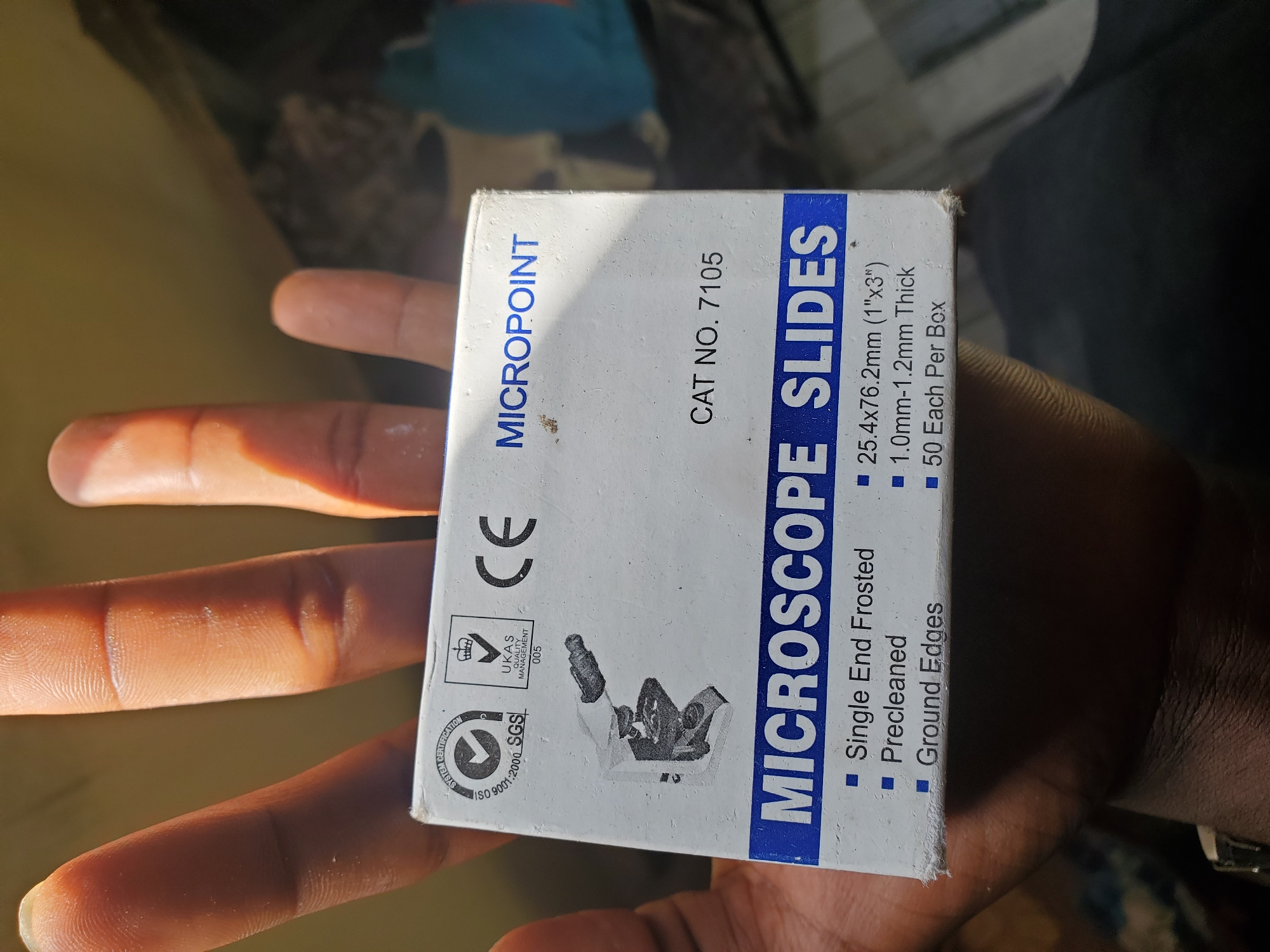 The image is a close-up shot of a hand holding a box of "MICROPOINT MICROSCOPE SLIDES." The box is white with blue lettering and features an illustration of a microscope, certification logos (SGS, UKAS), and the "CE" mark. Text on the box lists features such as "Single End Frosted," "Precleaned," "Ground Edges," dimensions "25.4x76.2mm (1"x3")," thickness "1.0mm-1.2mm," and "50 Each Per Box." The hand holding the box appears to be of dark complexion. The background is out of focus and suggests an indoor environment with muted colors. The image quality is sufficient to read the text on the box. The image is taken in Jalingo, Nigeria.