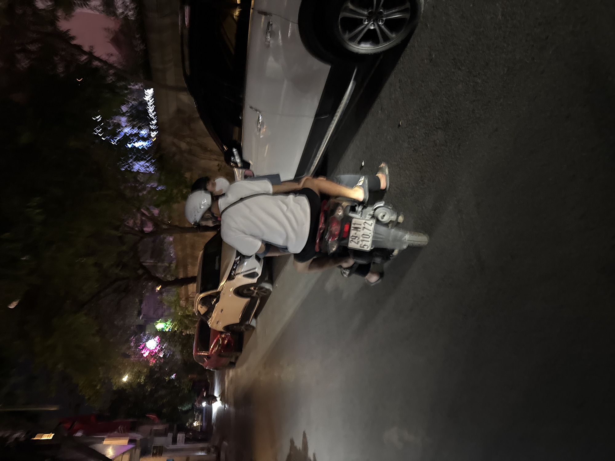 In this nighttime shot of a street in Hà Nội, Vietnam, a person wearing a white t-shirt, black shorts, and a white helmet is seated on a motorcycle. The rider is leaning forward, with their arms resting on their knees. The motorcycle is parked on the right side of the frame, next to a white car. The car's wheel is visible on the left side of the image. Further down the street, other vehicles, including a red car, are parked. The background shows trees and some colorful, blurred lights, suggesting a festive or well-lit area. The street appears wet, indicating recent rain or a damp surface. The license plate of the motorcycle reads "29-N1 510.72". The overall mood is subdued due to the nighttime setting, with the artificial lights creating a slightly surreal atmosphere.