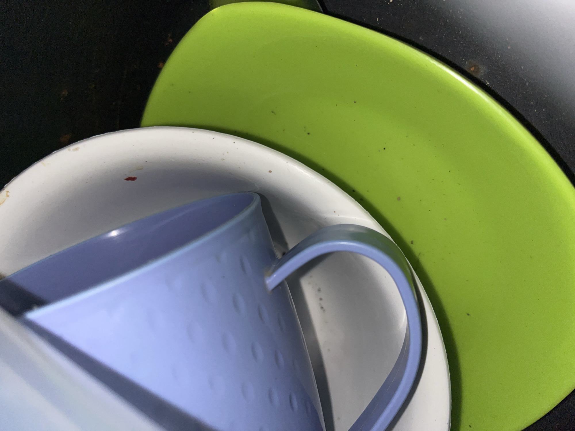 The image shows a close-up, angled view of a stack of dishes. The top item is a light purple mug, which is tilted and resting on a white plate. The white plate, in turn, is placed on a bright green plate. The background is dark and appears to be a countertop or sink area, with some small specks of debris visible on the surfaces of the dishes and in the background. The lighting is somewhat dim, creating shadows and highlighting the textures of the dishes. There are no people visible in the image. The location in the city of Benuaraja, Indonesia is not discernible from the visual information in the image.