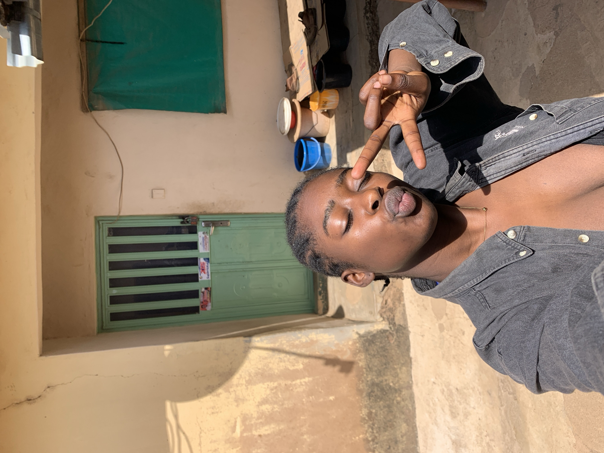 An individual is posing with a peace sign near their eye in an outdoor setting. The person is wearing a dark gray collared shirt. The background suggests a building exterior in Rafin Par, Nigeria, with a pale yellow wall, a green window covering, and a green door with dark horizontal bars. There are several buckets and containers visible on a higher level of the building. The foreground consists of a textured, light-colored surface, presumably the ground.