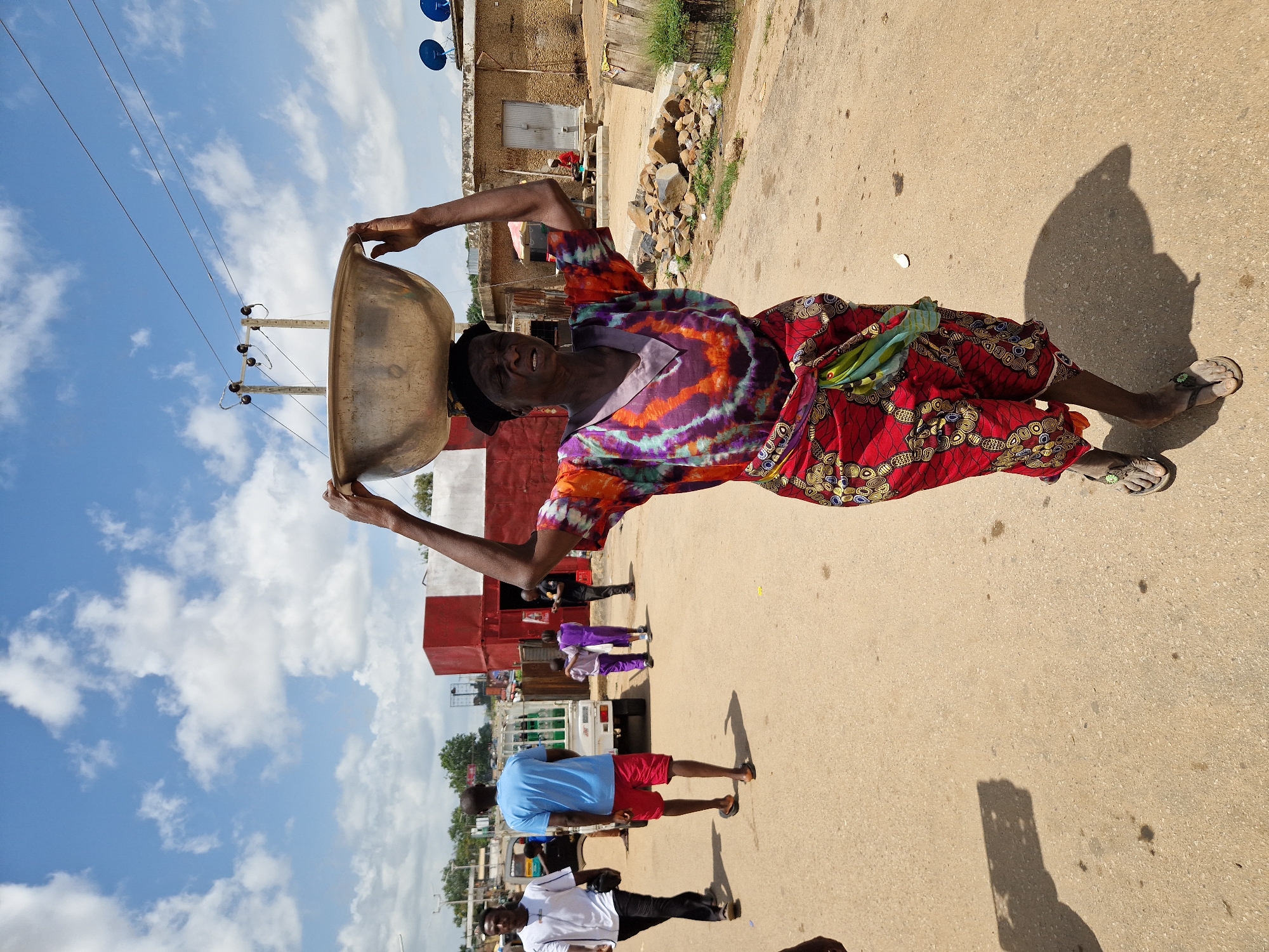 In this image, a woman carries a large metal bowl on her head, balancing it with her hands as she walks on a dirt road. She is wearing a colorful, tie-dye patterned shirt and a red and yellow patterned skirt, along with simple flip-flops. The scene appears to be a rural or semi-urban setting, with buildings visible in the background, some made of mud brick. Rocks are piled near one of the buildings, and there's some vegetation. Utility poles with wires are also present.
Several other people are visible in the background, walking or standing. One person in the foreground is wearing a blue t-shirt and red shorts. Another person in the far foreground wears a white t-shirt and black pants. The lighting suggests it is daytime, with a bright sky filled with scattered white clouds. The sun casts a distinct shadow of the woman with the bowl on the road. The overall impression is of a community going about its daily activities under a clear sky. There is no visible text in the image.