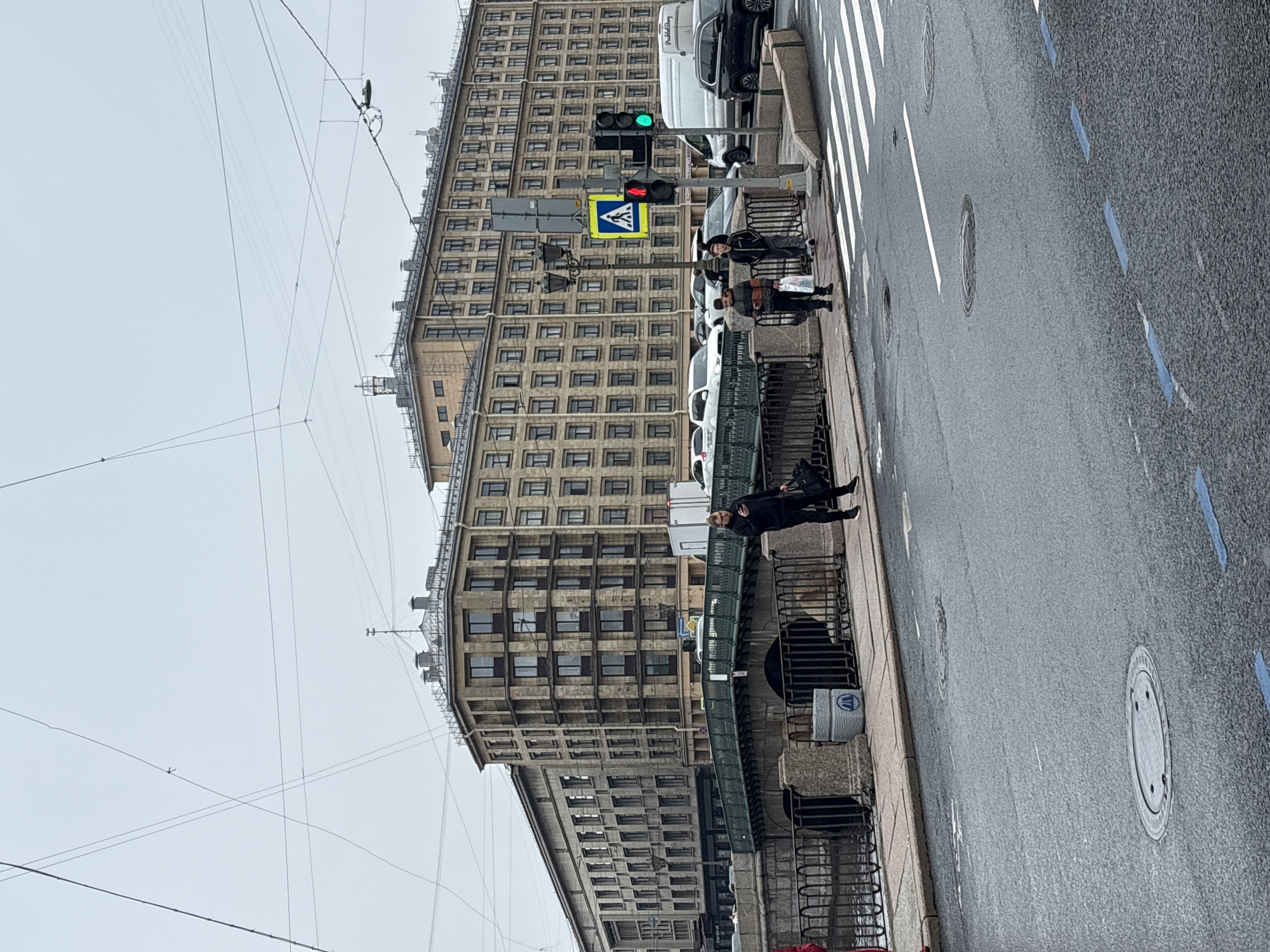 The image depicts a street scene in Saint Petersburg, Russia. A tall, Soviet-era building with numerous windows dominates the background. In the foreground, a sidewalk runs along the street, separated by a dark metal fence. People are present, either sitting on a low structure or standing along the sidewalk. A crosswalk with white stripes is visible on the street. Traffic lights display a green signal, and a pedestrian crossing sign is present. Overhead, wires crisscross the sky. The street is wet, indicating possible recent rain. Manhole covers are visible on the road surface. The overall weather appears overcast and gray.