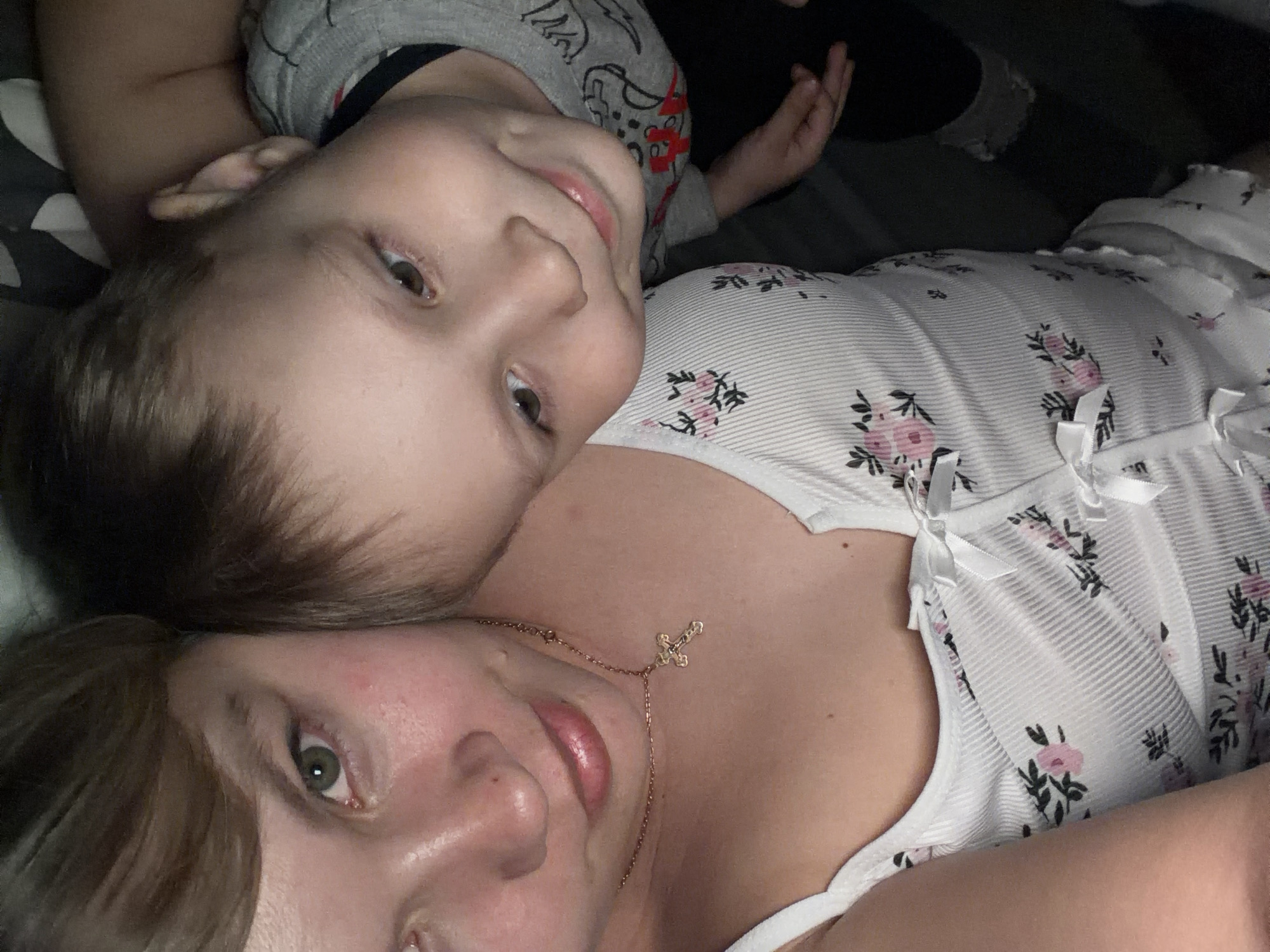A woman and a young child are lying down. The child, wearing a gray patterned shirt, is positioned with his head near the woman's face. The woman is wearing a white floral top with spaghetti straps and a gold necklace with a cross pendant. They are in an indoor environment. There are no obvious indicators in the image to pinpoint the exact location in Chaikovsky, Russia.