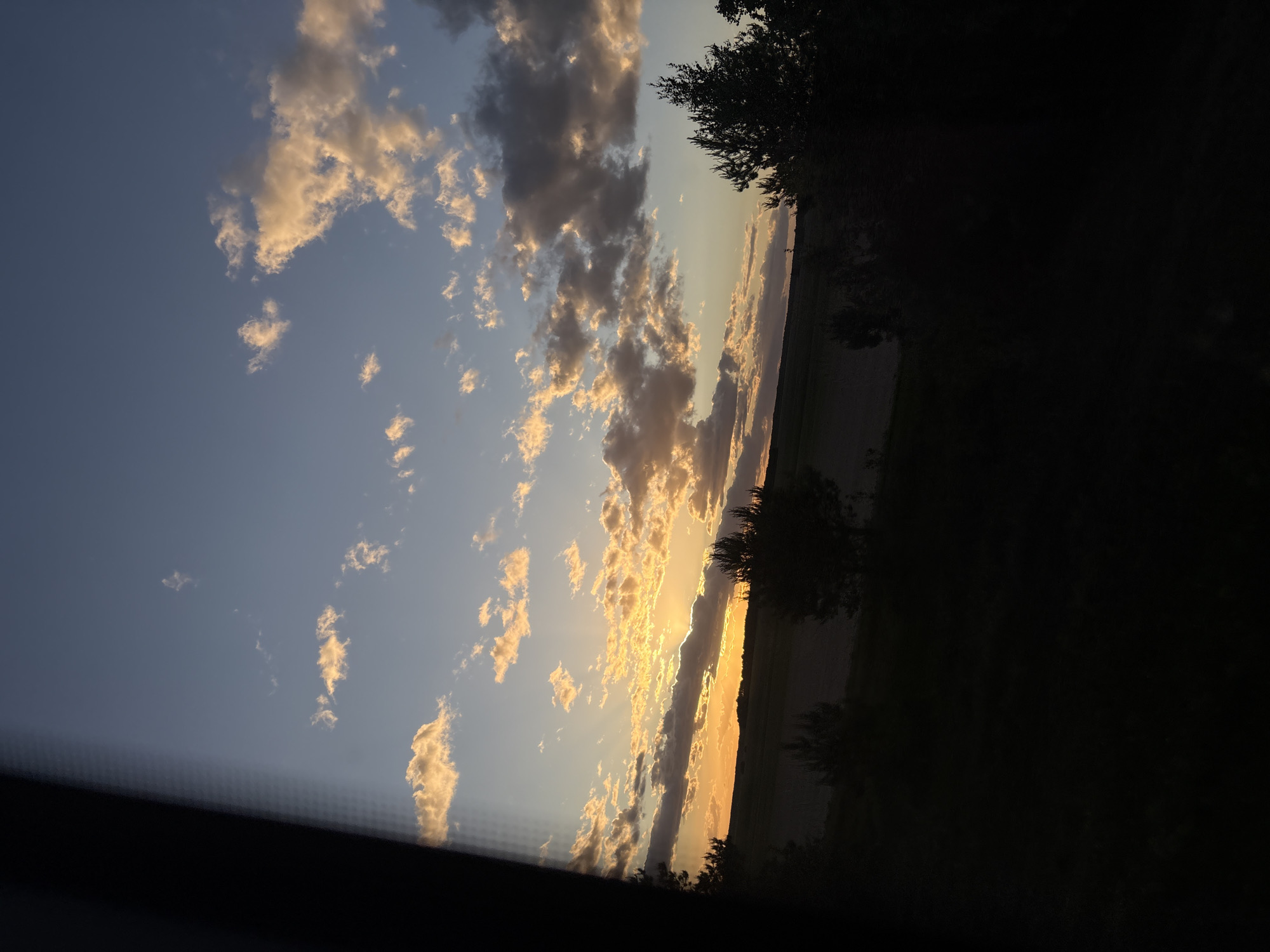The image shows a sunset in Мажэйкаўскі сельскі Савет, Belarus. The sky is a gradient of blue to orange with scattered cumulus clouds catching the golden light of the setting sun. Silhouettes of trees and vegetation are visible along the horizon line. The foreground is mostly dark.