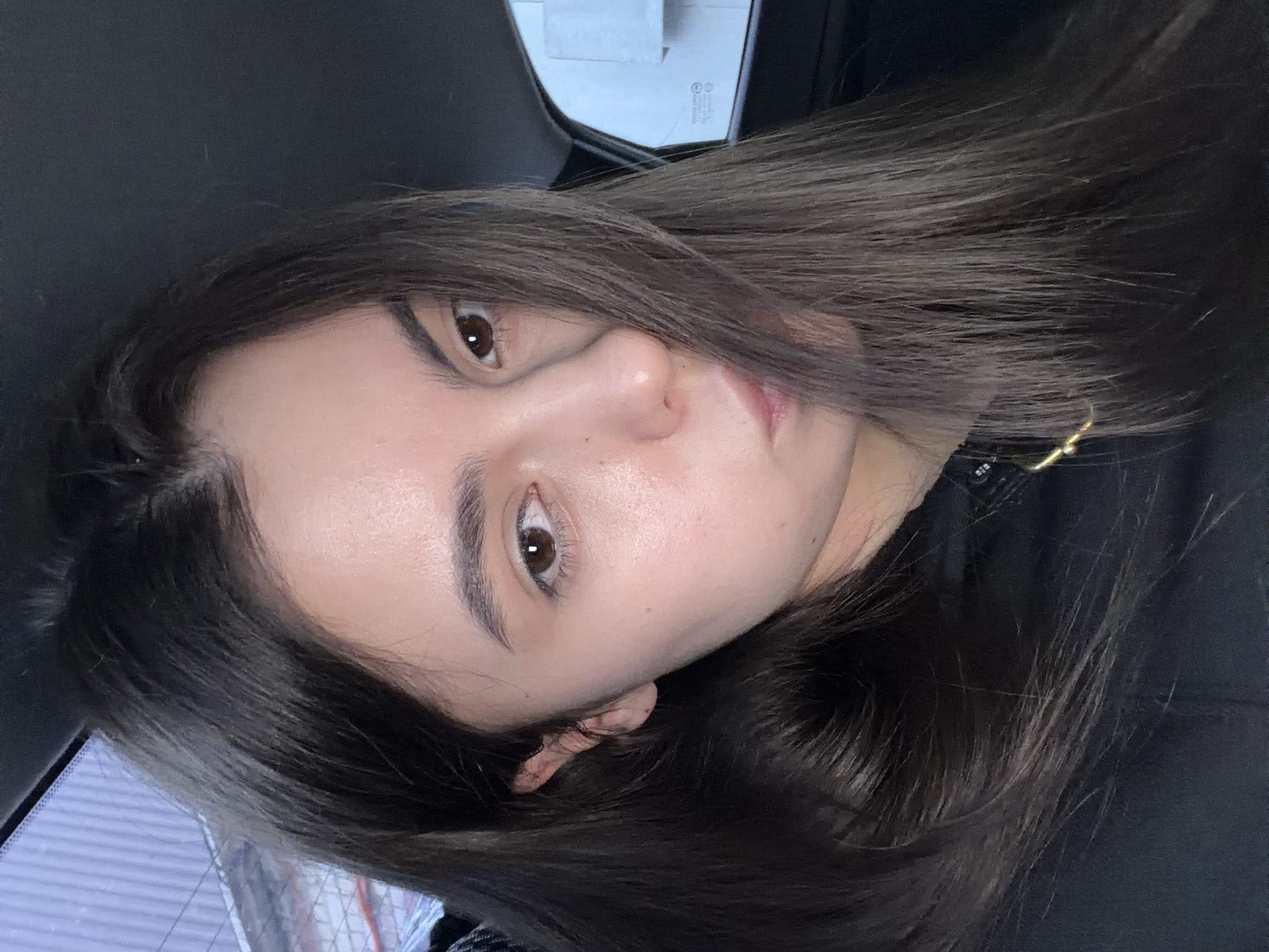A close-up, slightly angled portrait of a woman with long, dark brown hair. She has fair skin, brown eyes, and arched eyebrows. She is wearing a black top and a gold necklace. The background is blurred and appears to be the interior of a car, with a window and a glimpse of what may be a building visible. There is no evidence to suggest that this image was taken in Chaikovsky, Russia.