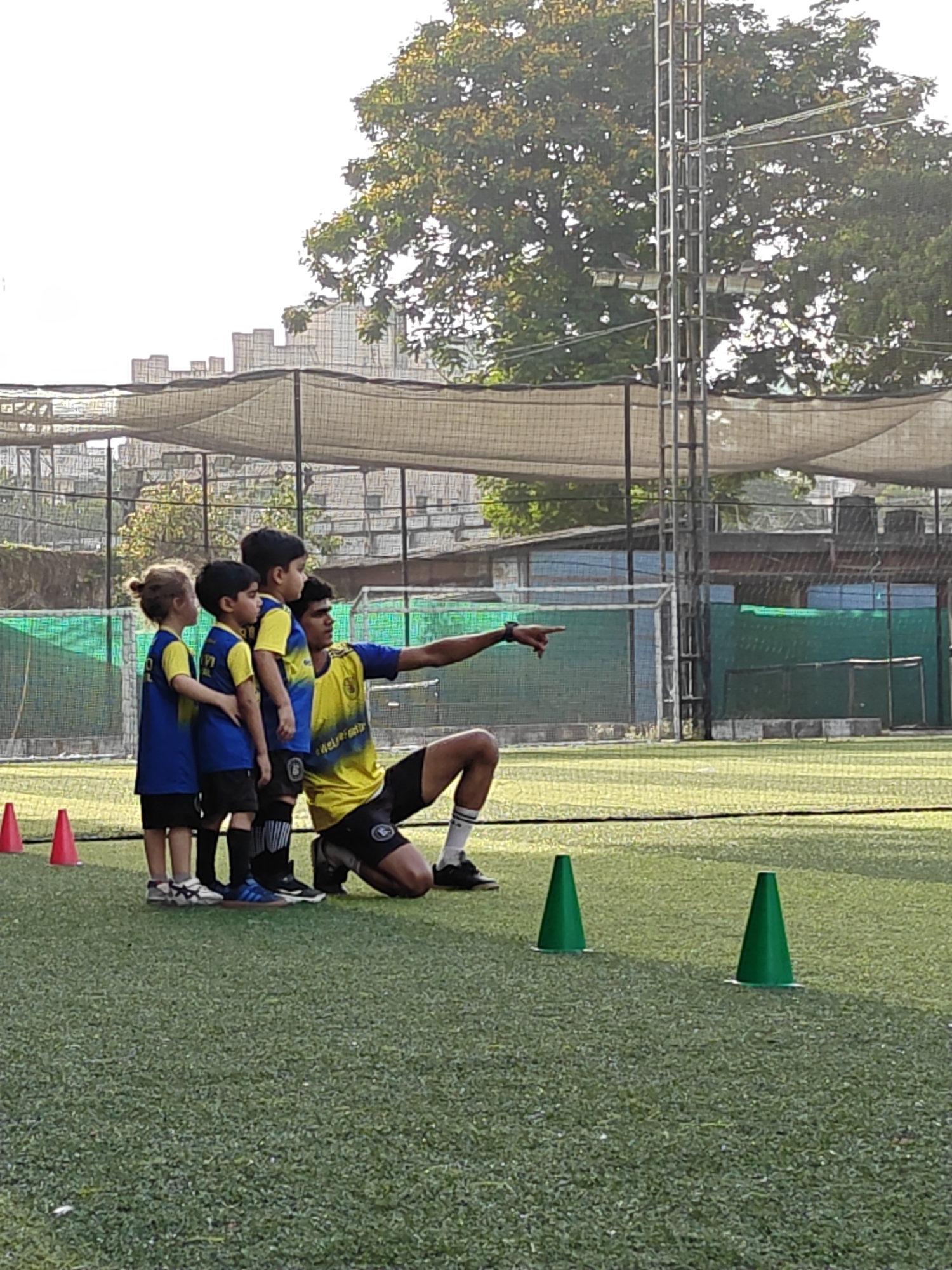 A soccer coach, in a blue and yellow team uniform, is kneeling on a green soccer field with three young children standing next to him in matching team uniforms. The coach is pointing with his right arm towards an area outside of the frame of the photo. Three green cones are arranged in front of the coach and his students. Three red cones are to the left of the group. The setting of the photo is in an outdoor stadium, complete with a net canopy, surrounding walls, and a metal light tower with trees in the background. The location is likely Mumbai, India.
