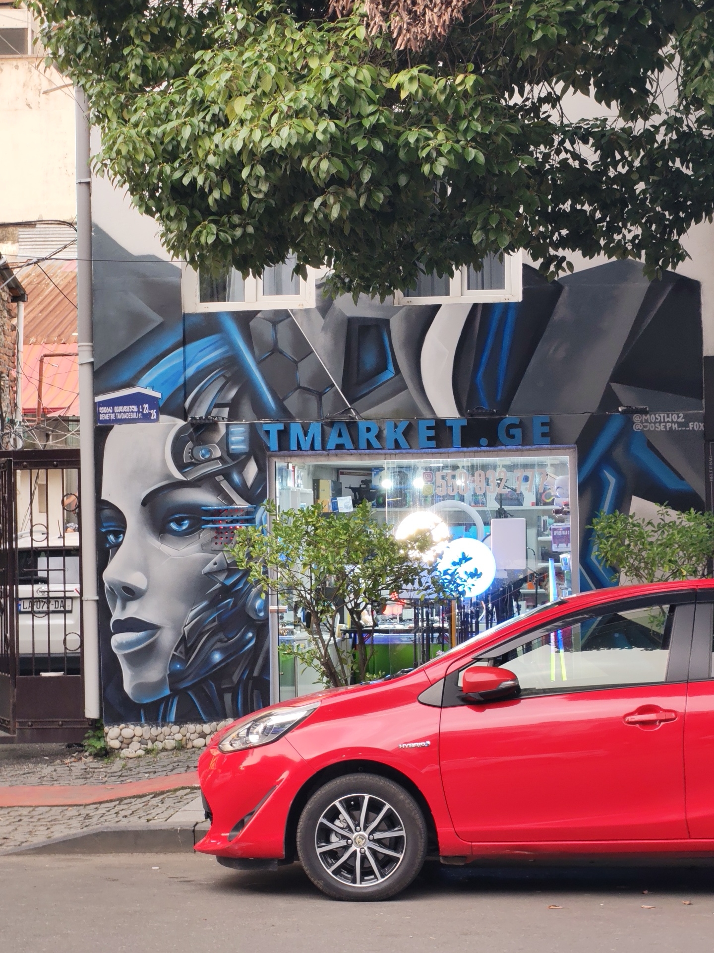 A red car is parked on a street in Batumi, Georgia, in front of a building with a mural on its facade. The mural depicts a stylized, futuristic face with blue accents and metallic elements, possibly representing a robot or android. Above the mural, there is a sign for "TMARKET.GE" with blue lettering. The building appears to be a shop or business, with a display window showing various items. Trees and foliage partially obscure the building and mural. The street is paved with cobblestones, and a sidewalk is visible. The license plate on a white vehicle partially visible behind a gate on the left side of the image reads "LA 077 DA". The overall scene suggests an urban environment in Batumi.