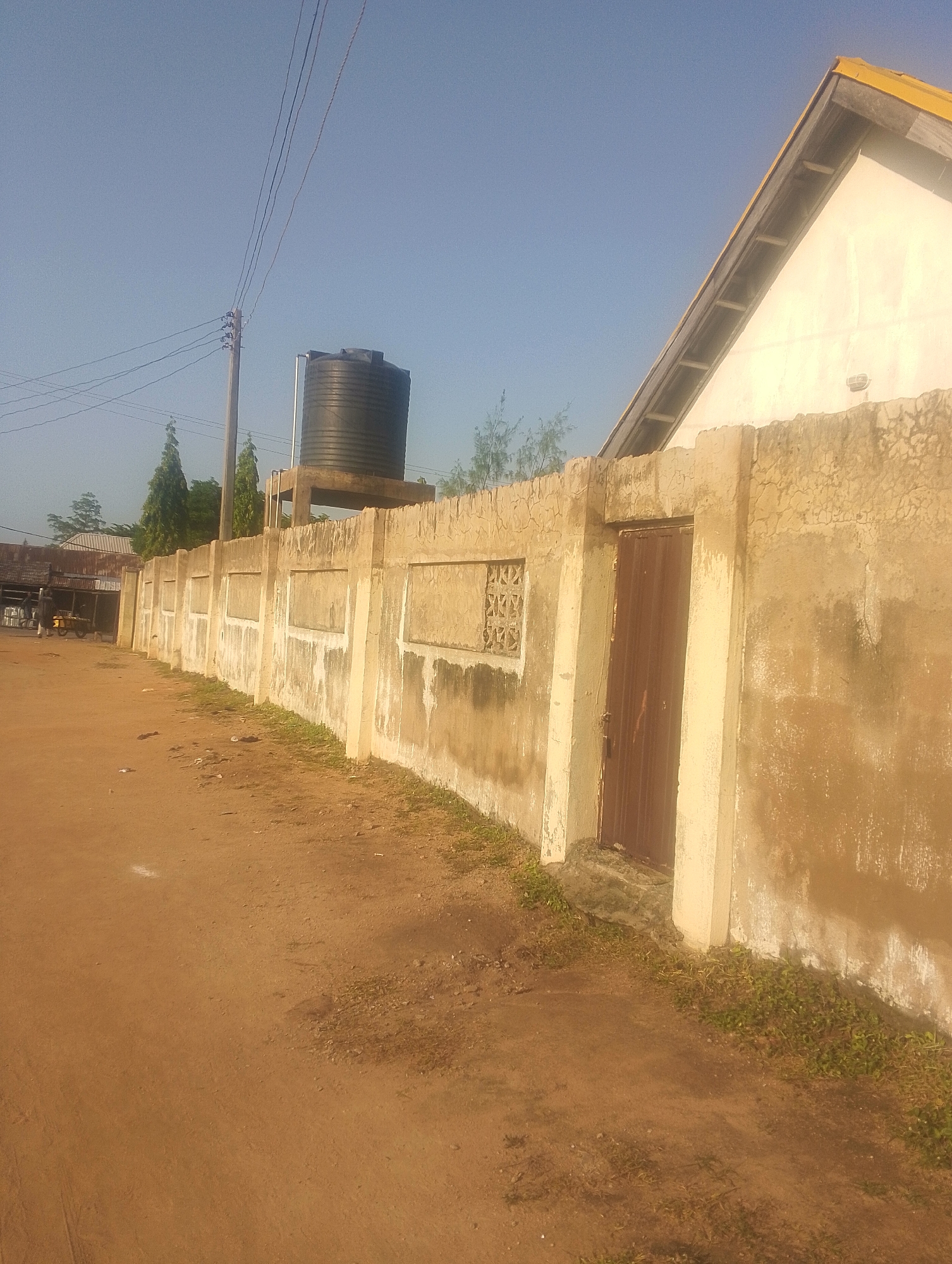 The image depicts an outdoor scene on a dirt road in Jalingo, Nigeria. A long, weathered, concrete wall runs along the right side of the frame, showing signs of age and discoloration. Embedded within the wall are decorative rectangular panels, and at one point, a metal door with vertical slats is visible. To the left of the wall, a dirt path leads away from the viewer, with sparse, dry vegetation growing along the base of the wall. In the background, beyond the wall, a portion of a building with a yellow-tiled roof and white walls can be seen, along with a large black water tank supported by a concrete structure. Electrical wires on wooden poles are strung across the sky, suggesting the presence of utilities. In the far left background, a glimpse of other buildings and some trees is visible. The sky is clear and blue, indicating a sunny day. No people are clearly identifiable in the scene.