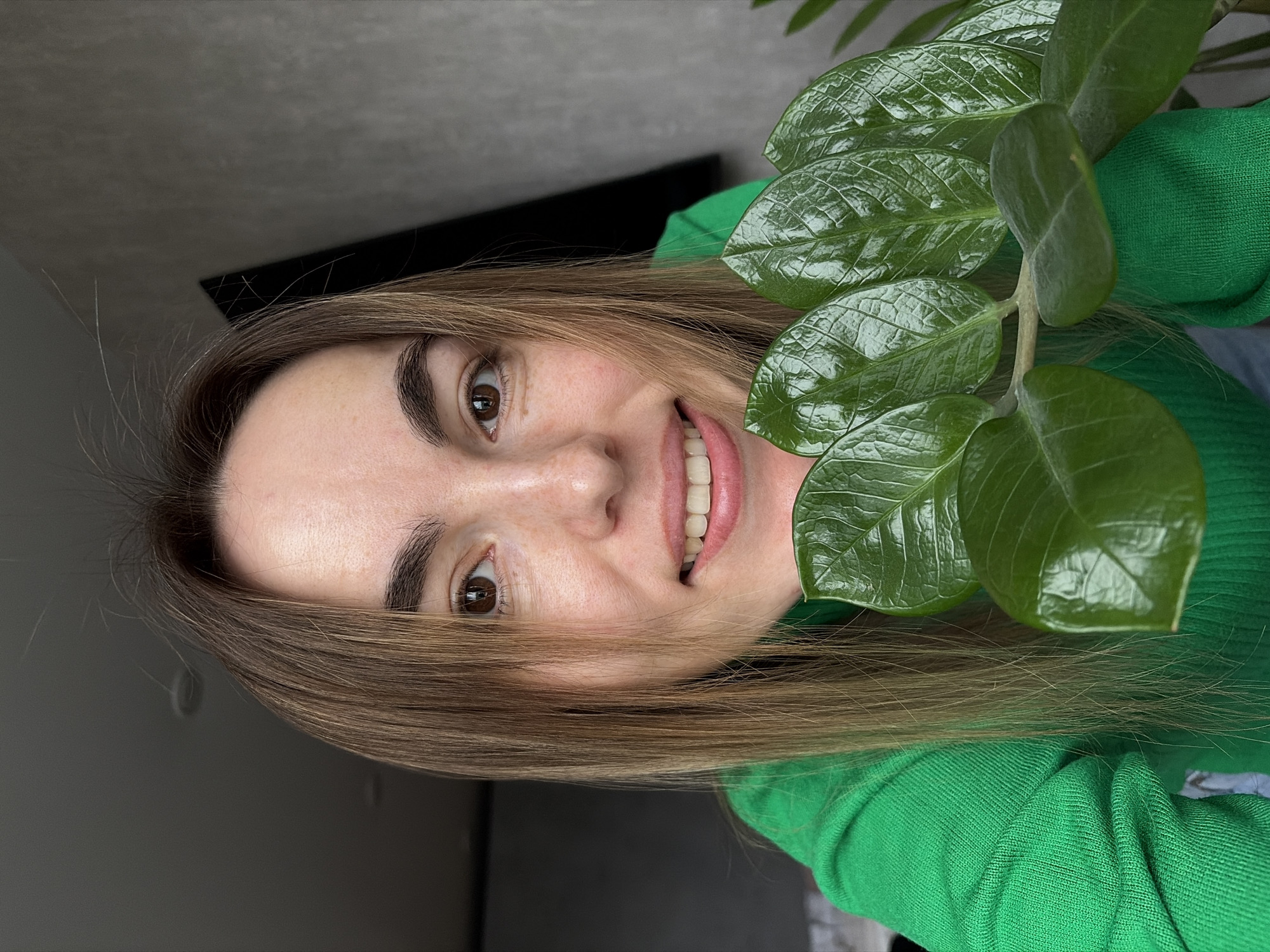 A woman with brown hair and brown eyes is smiling at the camera, partially obscured by the green leaves of a plant. She is wearing a green sweater. The background is a plain grey wall with a black object mounted on it. There is no indication of the city of Izhevsk, Russia.