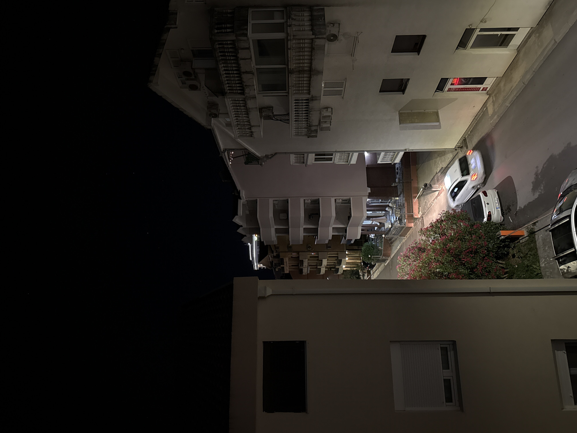 This is a nighttime, high-angle, wide shot of a street in Budva, Montenegro. The scene is dominated by buildings and a road. On the left, a light-colored building with a dark rectangular window is visible. To the right of this building, the street curves, lined with multi-story buildings. The buildings have balconies, windows, and air conditioning units. The street itself is paved and illuminated by artificial lights. Two white cars are on the street, one is a hatchback in motion, and the other is parked. A flowering bush with pink flowers is also visible. The sky above is dark, with a few faint stars.