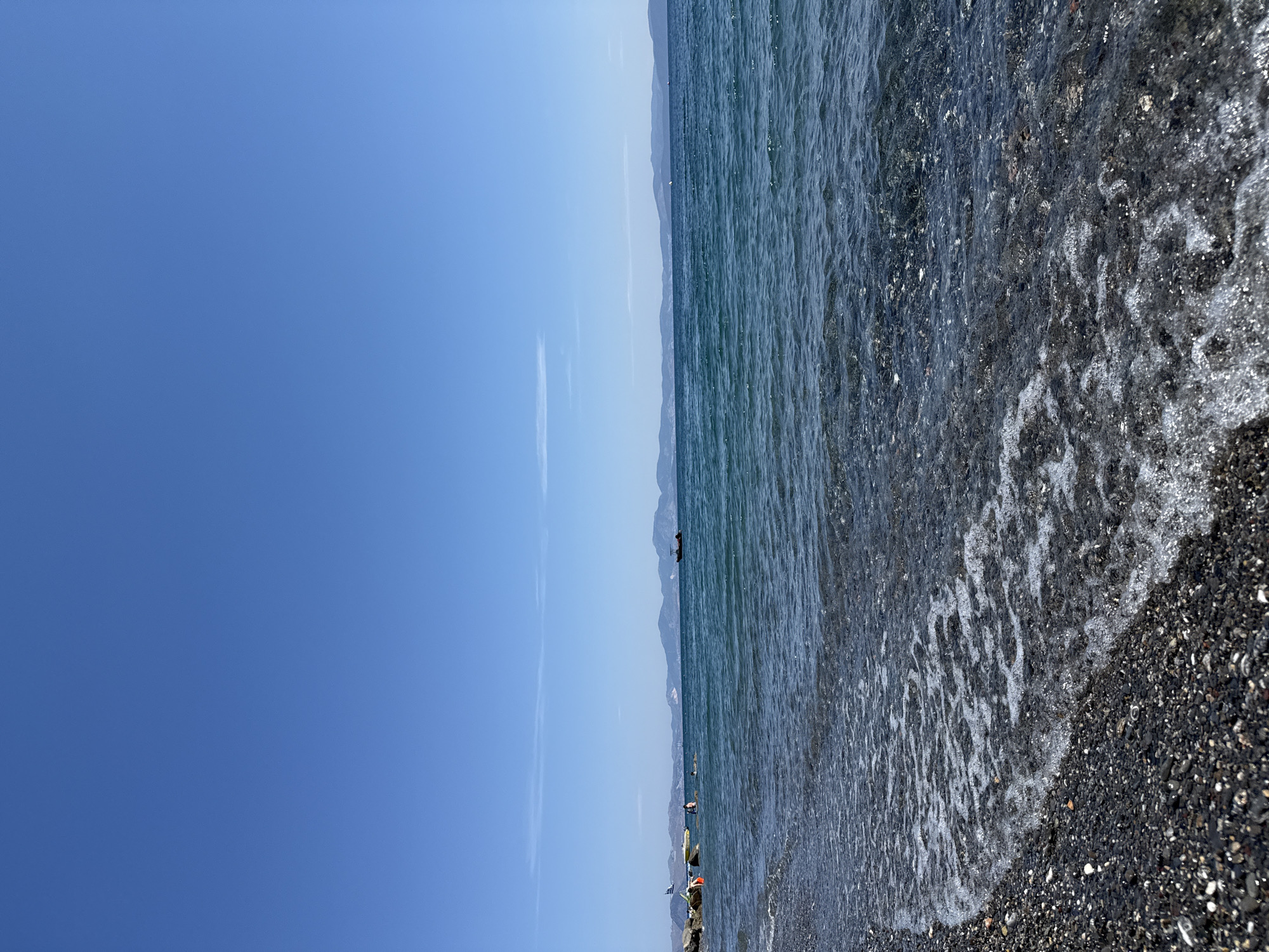 The image depicts a beach scene in Kos, Greece. The beach has dark-colored pebbles. The water is clear with visible waves. The sky is blue. In the distance, there are mountains. There are a few people visible in the water and on the shore. There are also some unidentified objects on the beach. The orientation of the photograph is horizontal.