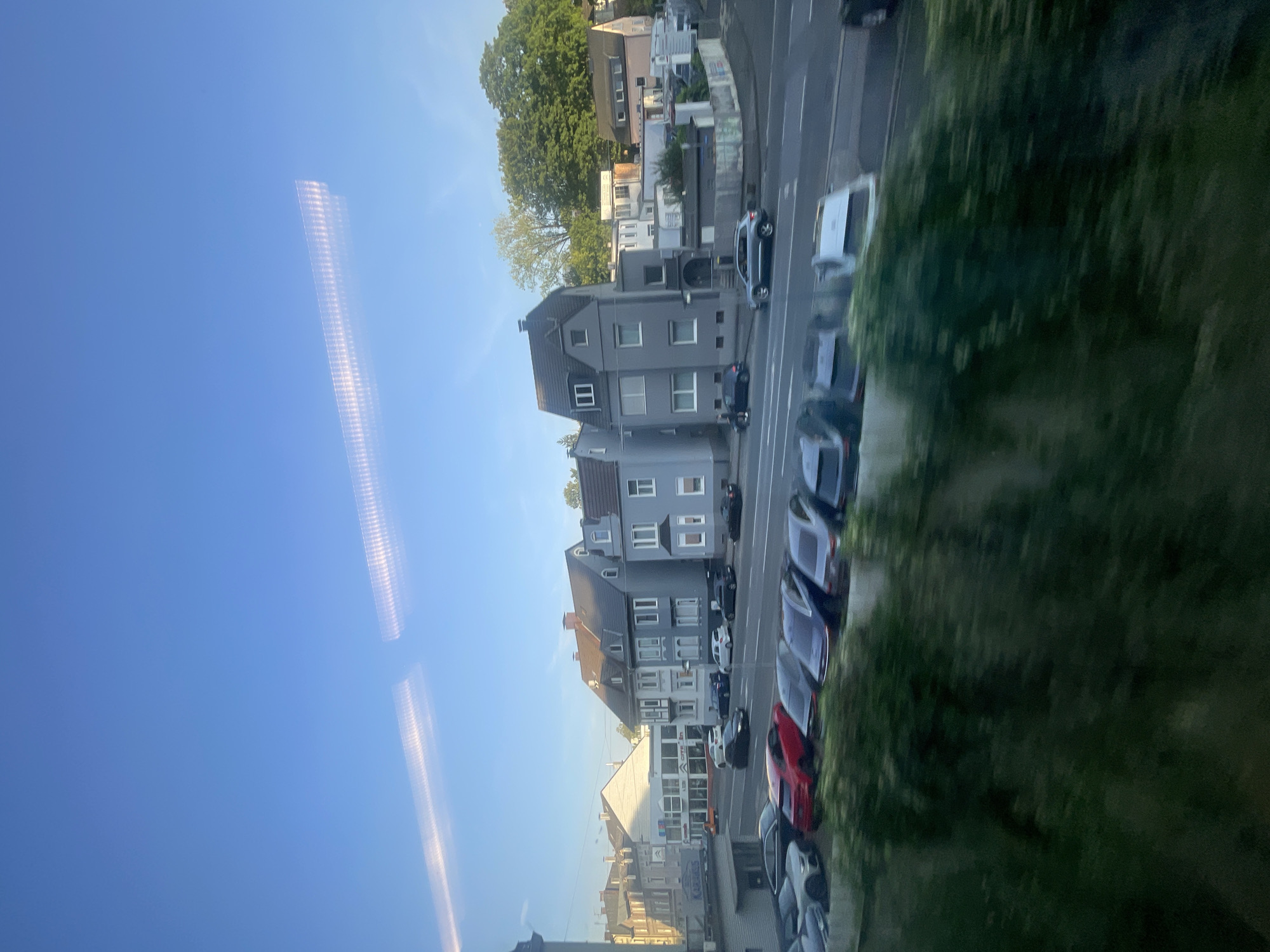 The image captures a street view in Iserlohn, Germany, with several residential buildings and a row of parked cars. The sky is a clear blue with two horizontal streaks of light, possibly from the sun reflecting off a window or passing train. In the foreground, a line of vehicles, including a red car and several SUVs and sedans, are parked along the street. Behind the cars, a row of multi-story buildings, primarily gray and white, are visible. One building has a prominent gray facade with dark gray roofing and multiple windows. Further down the street, more buildings, including a lighter-colored one with an arched entrance, are visible. Trees and greenery are present, particularly on the right side of the image, adding to the urban residential setting. The overall impression is that of a daytime scene in a European town.