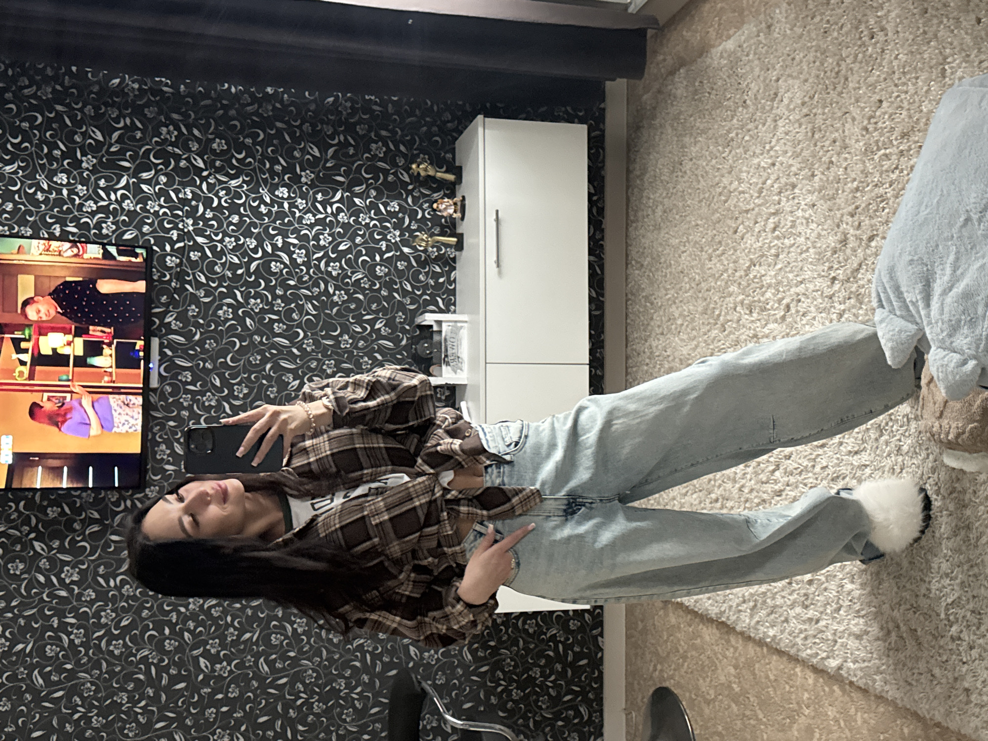 A woman is taking a selfie in a room with floral wallpaper, a TV, and a white cabinet in what is likely a residential area. The room is furnished with a beige rug. The woman is wearing jeans and a flannel shirt. She is holding a smartphone in her hand and taking a picture of herself. The television on the wall is on, and its screen displays various colors and shapes. Based on the available information, determining the exact location in the city of Chaikovsky, Russia is not possible. The description focuses on the details visible in the image.