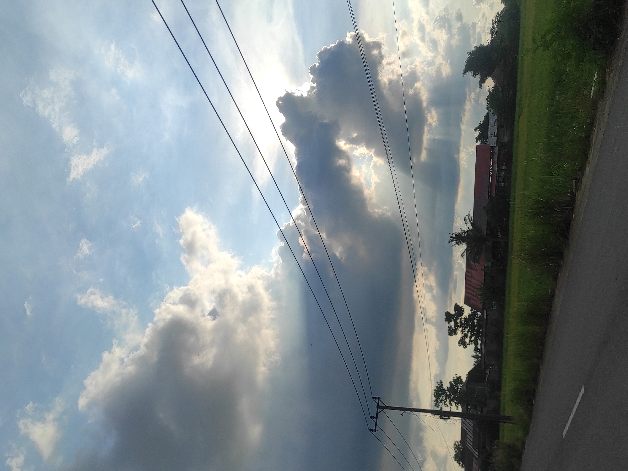 The image shows a view of the sky with clouds and sunlight, taken in Lhokseumawe, Indonesia. Power lines are prominently featured in the foreground, running diagonally across the frame. A utility pole is visible at the lower portion of the image. To the right, a road is adjacent to a grassy area and some buildings.