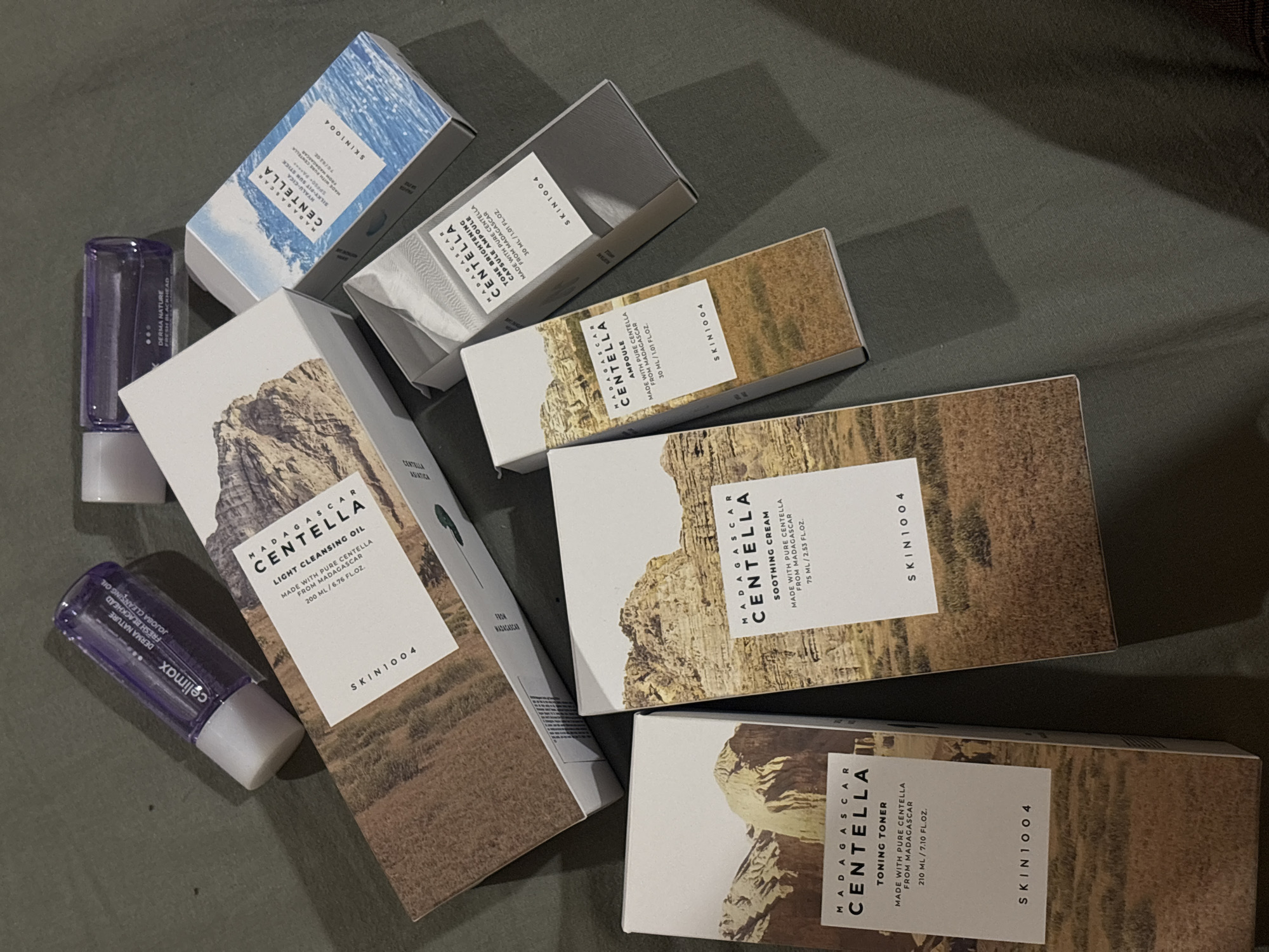 The image shows a collection of skincare products from the brand SKIN1004's Madagascar Centella line, laid out on a surface, likely a table or floor.
The products include:
* Madagascar Centella Toning Toner (210 ml)
* Madagascar Centella Light Cleansing Oil (200 ml)
* Madagascar Centella Soothing Cream (75 ml)
* Madagascar Centella Ampoule (30 ml)
* Madagascar Centella Tone Brightening Capsule Ampoule (30 ml)
* Madagascar Centella Hyalu-Cica Silky-Fit Sun Stick
* Celimax Derma Nature Fresh Blackhead Jojoba Cleansing Oil
All product boxes share a common design featuring a landscape image on the front and the SKIN1004 logo. The blackhead cleanser is not part of the SKIN1004 line. The environment appears to be indoors.
The image does not depict any location in Bečići, Montenegro.