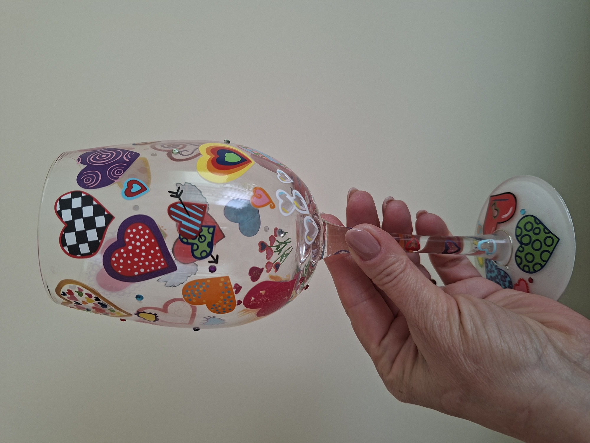 A close-up, side-view shot captures a hand holding a decorative wine glass. The glass is clear and adorned with a colorful array of heart-shaped designs, featuring various patterns such as polka dots, checkerboards, and concentric circles. Some hearts have arrows passing through them, and there are also small floral embellishments. The hand, belonging to a person with light skin and neatly manicured nails, grips the stem of the glass, with the fingers wrapped around it. The background is a plain, light-colored wall, suggesting an indoor setting. The lighting appears to be neutral, without strong shadows, indicating it could be daytime or well-lit indoor conditions. No text is visible in the image. The scene is static, focused on the object and the hand interacting with it, evoking a sense of personal possession or appreciation for the decorative item. The visual focus is on the intricate details and bright colors of the hearts painted on the glass.