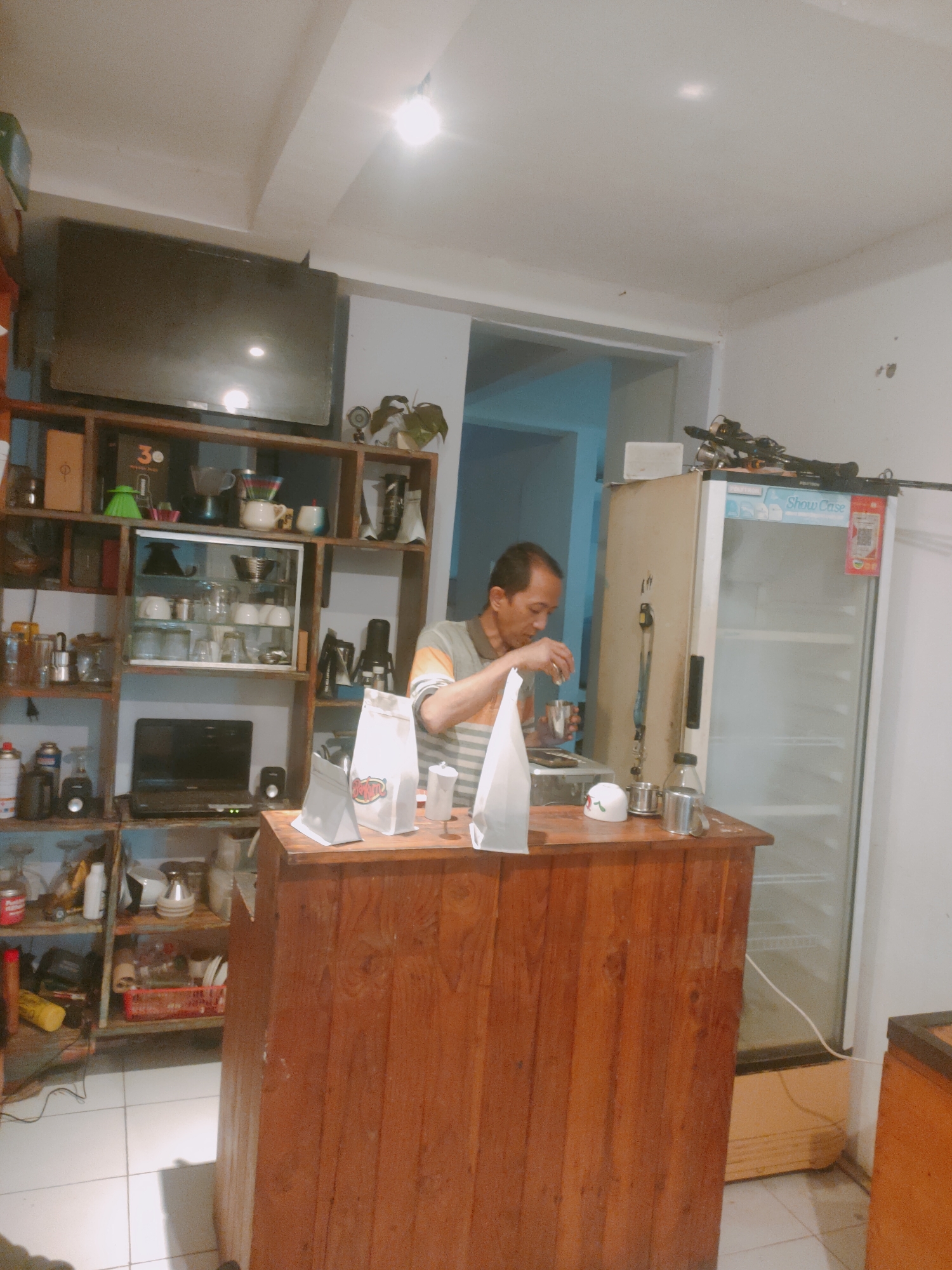 The image shows a man standing behind a wooden counter in what appears to be a small cafe or shop. The man, wearing a striped shirt, is in the process of making a drink, holding a small cup and a silver container. On the counter are several items including paper bags, a small white cup with a red design, and other containers. To the right of the counter is a large, clear-doored refrigerator. Behind the counter, a wooden shelving unit is filled with various items, including glasses, cups, bottles, and a laptop. A television is mounted on the wall above the shelves. The floor is tiled. The lighting appears to be artificial, coming from overhead fixtures. The overall impression is of a functional, albeit somewhat cluttered, commercial space. The location is identified as Sooko, Indonesia.