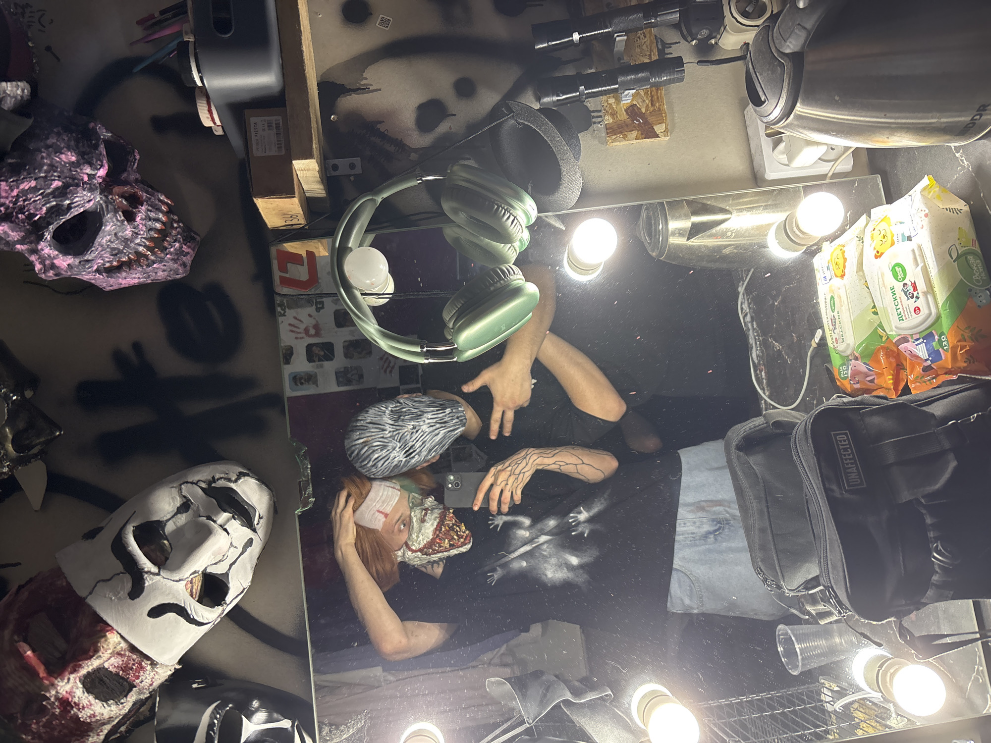 The image depicts a cluttered vanity surface. A person is reflected in the mirror, wearing a mask and head covering, and holding a phone up to take a picture. The person's reflection includes tattoos on their arm. The vanity is illuminated by multiple light bulbs.
Various items are scattered across the surface, including a pair of green headphones, two other masks, flashlights, a kettle, a pack of baby wipes, and a black bag labeled "UNAFFECTED."
The scene appears to be indoors, but there is no indication of the city of Saratov, Russia.